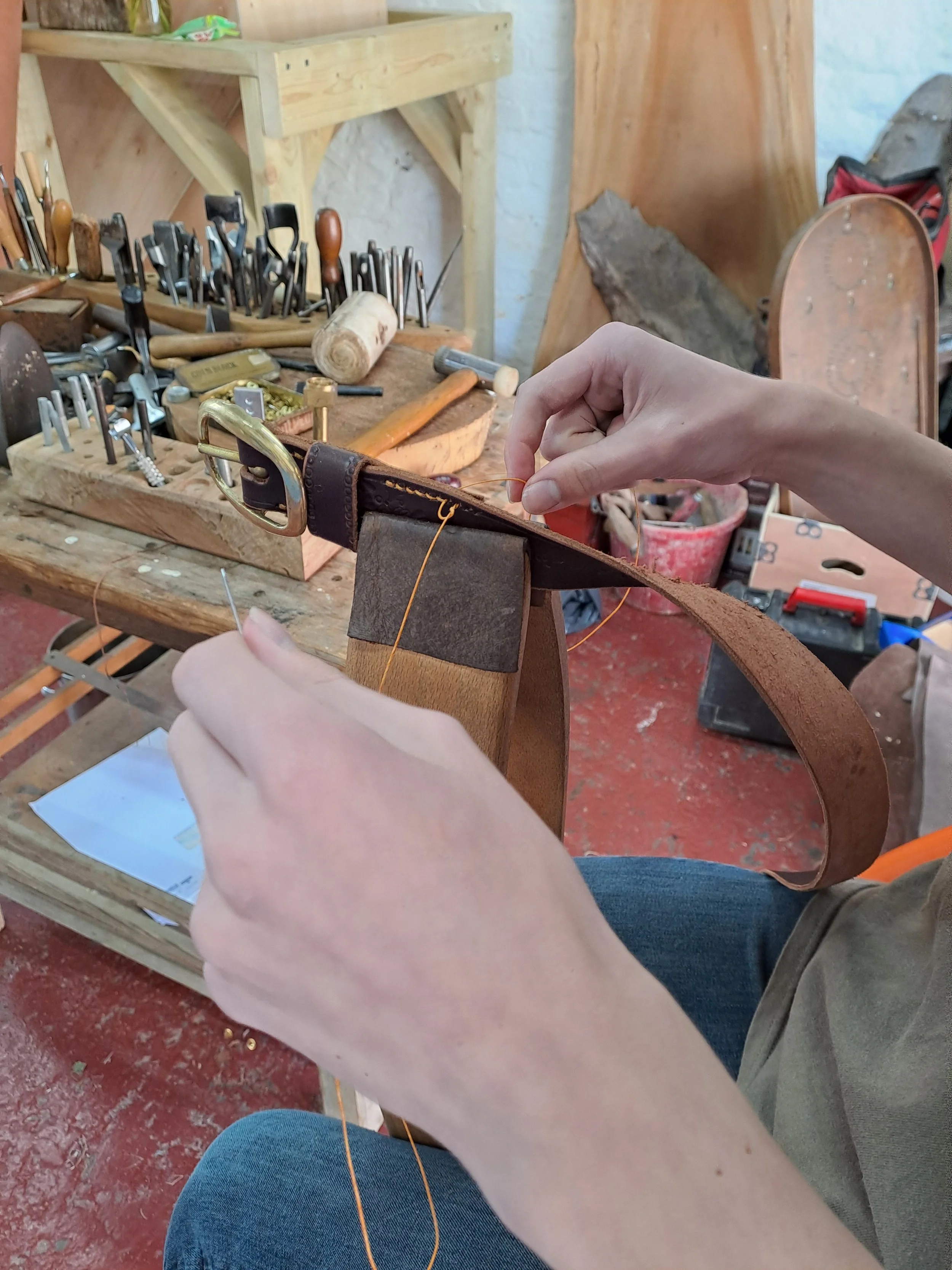 Person sewing a leather strap on a wooden shoe in a woodworking workshop with tools and wood pieces in the background.