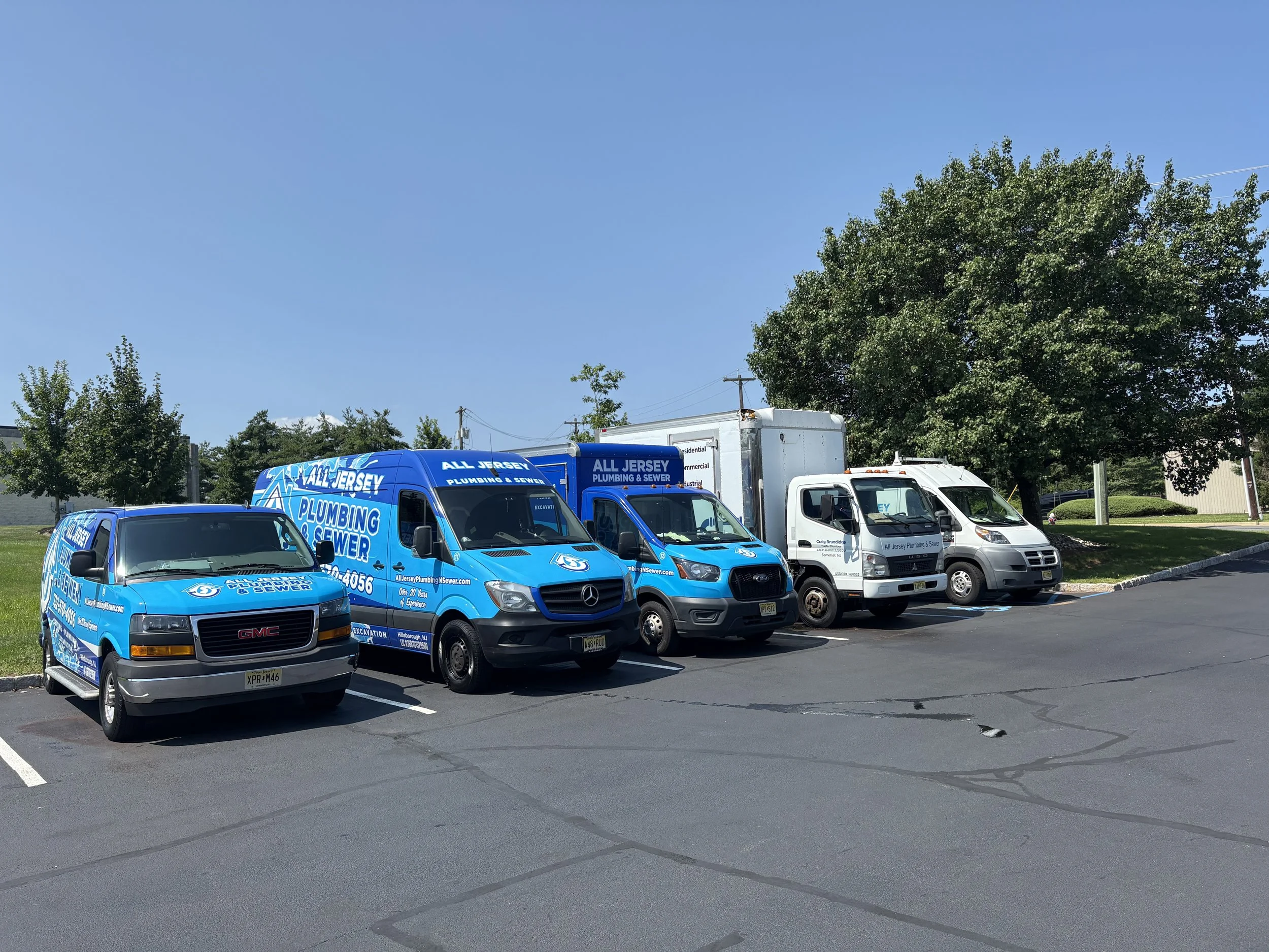 A row of five parked delivery service and plumbing vehicles on a paved parking lot, with trees and blue sky in the background. The first two vehicles are blue with company branding for All Jersey Plumbing & Sewer. The remaining three vehicles are white and silver with similar branding.