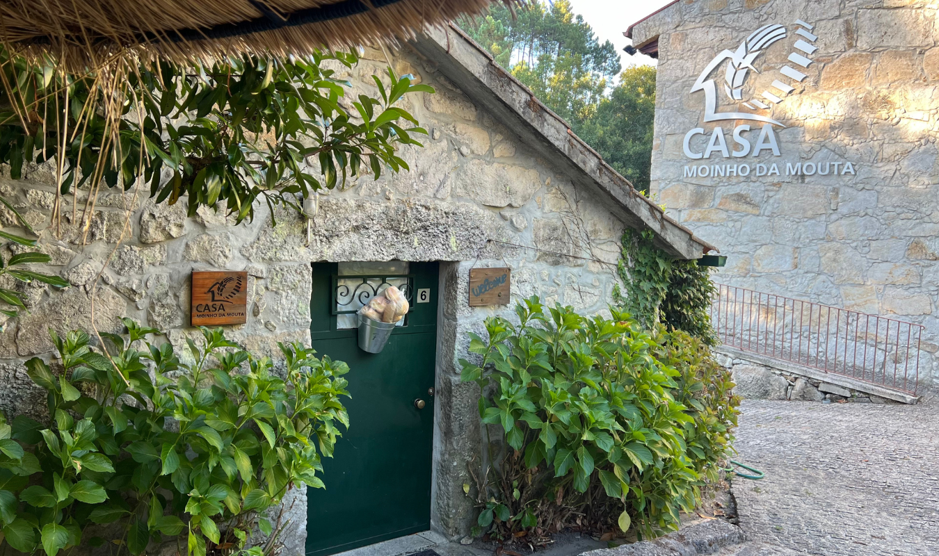 Edificação de pedra com porta verde, cercada por arbustos verdes, com placas indicativas, uma delas com a palavra 'Welcome', e uma grande placa na parede de pedra com 'CASA MOINHO DA MOUTA'.