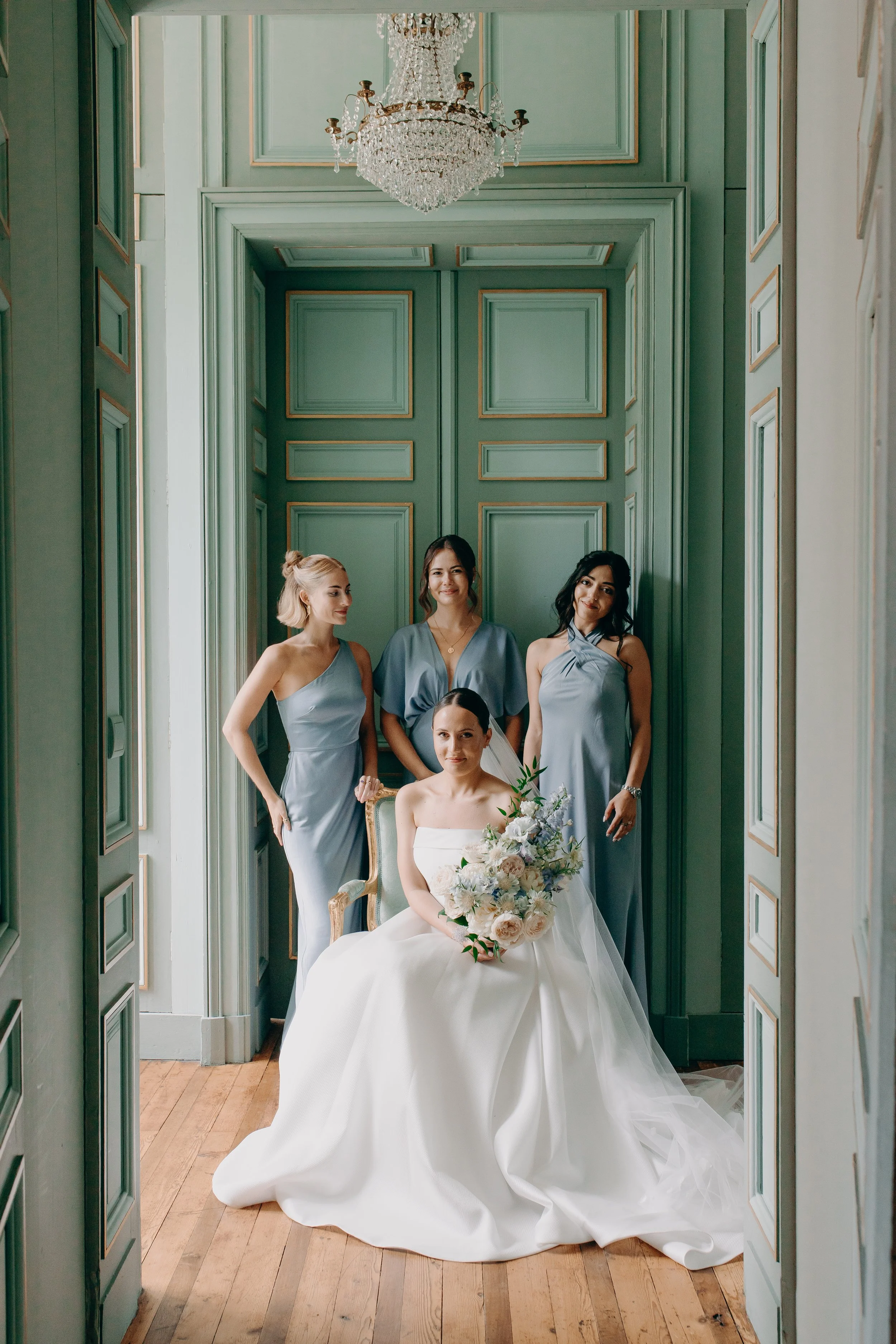 Bride in a white wedding dress seated with three bridesmaids in light blue dresses standing beside her, in a room with green paneled walls and a chandelier.