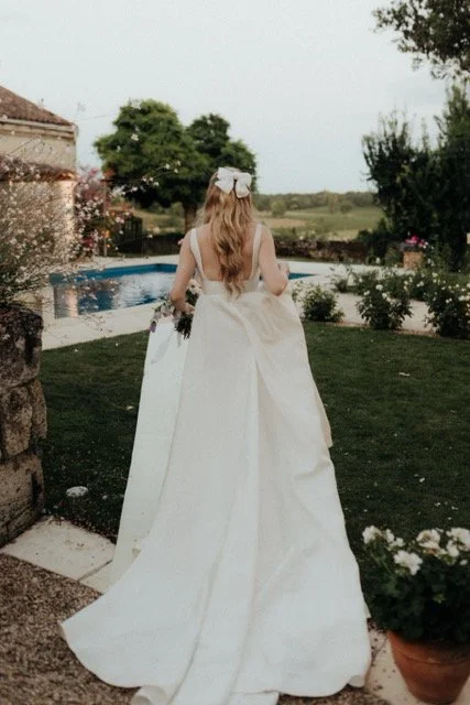 A bride in a white gown with a long train and a hair bow walking outdoors near a swimming pool.