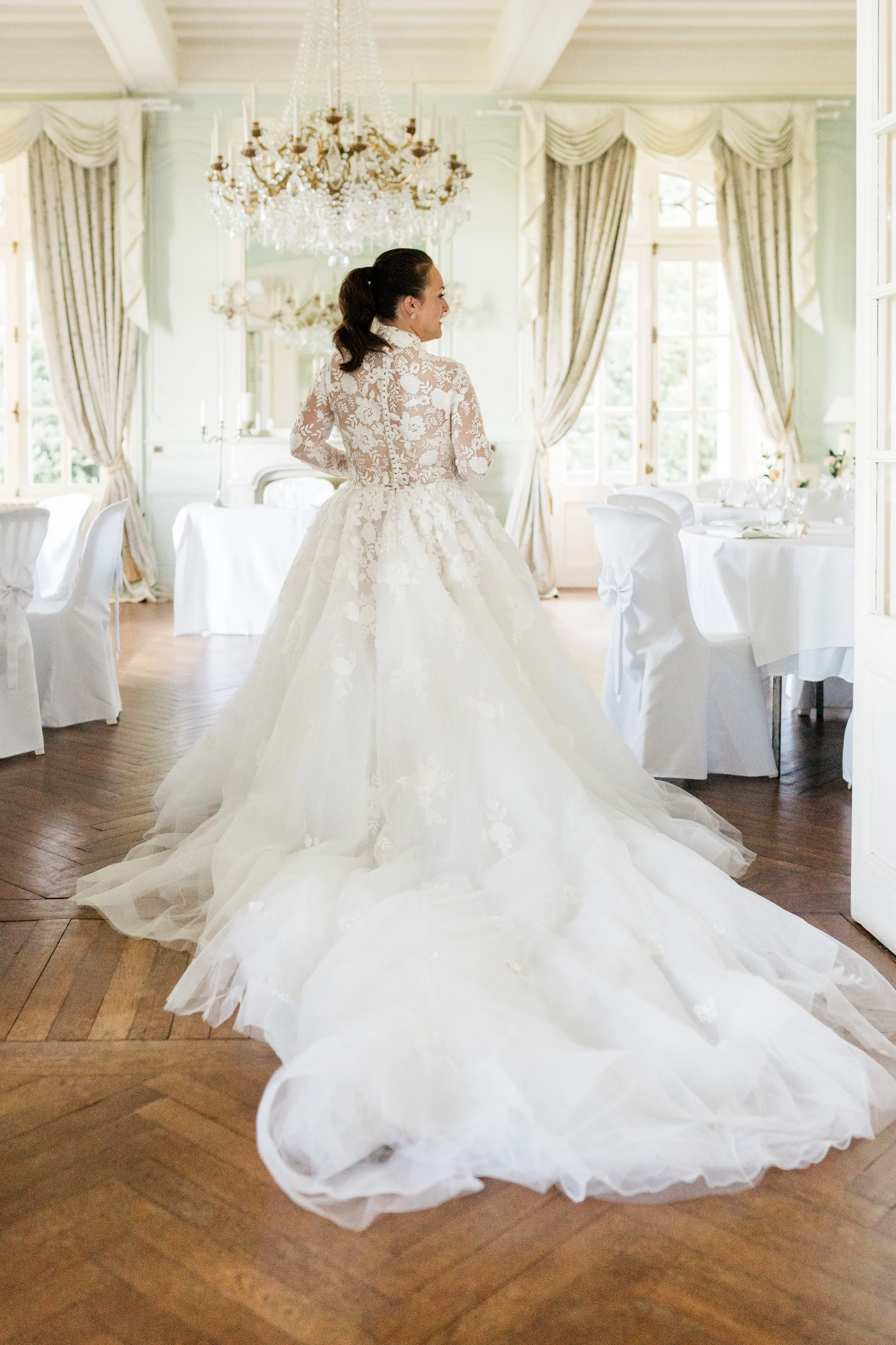 Bride in white lace wedding dress with long train stands in elegant room with chandeliers and draped curtains.