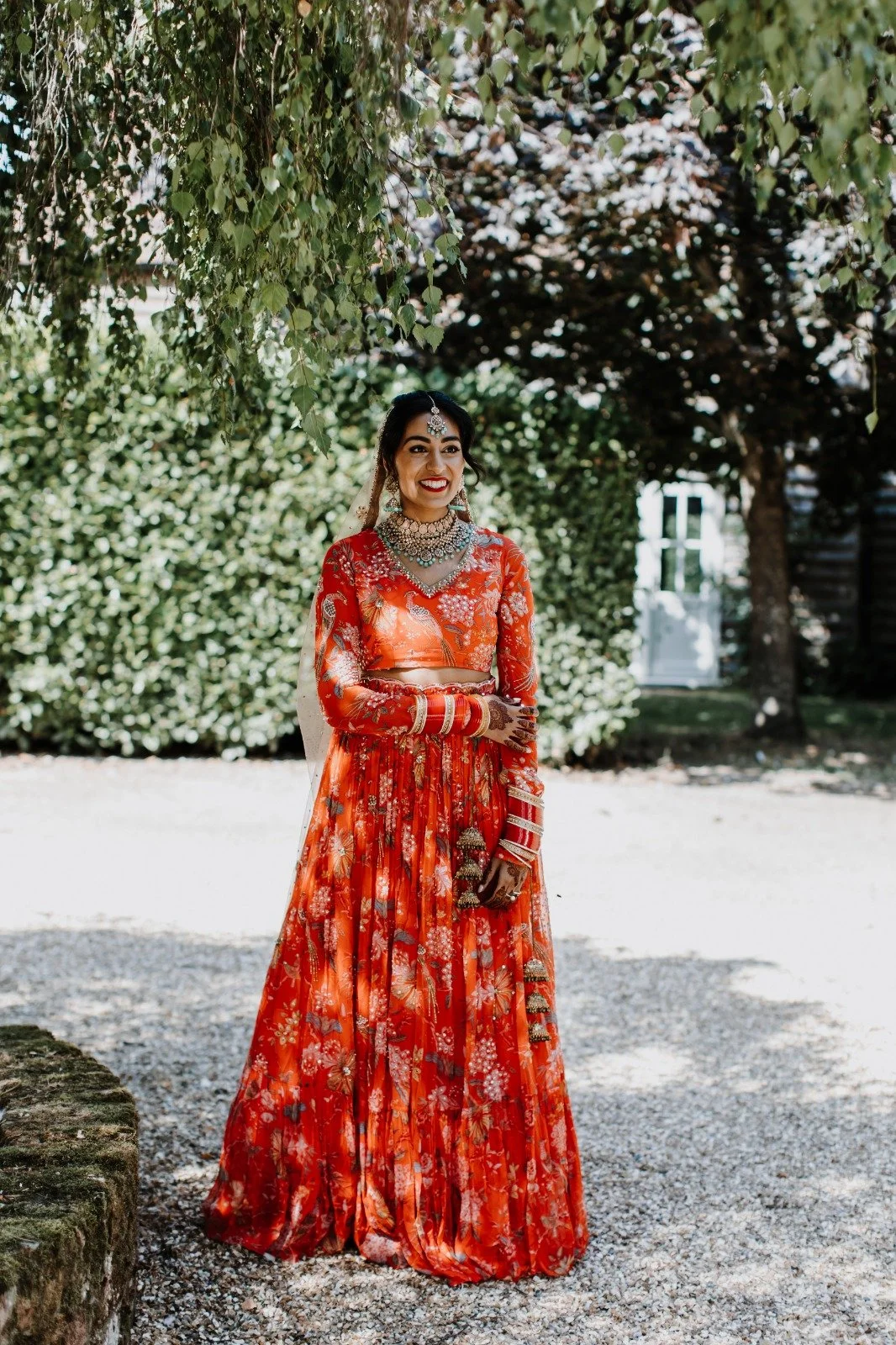 Woman in a red and gold traditional Indian attire, adorned with jewelry, standing on a gravel path under green leafy trees.