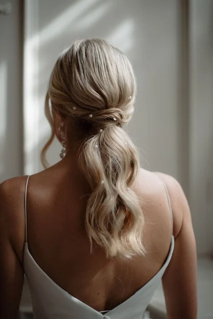 Woman with blonde hair styled in a ponytail with pearls, wearing a white sleeveless dress, seen from the back.