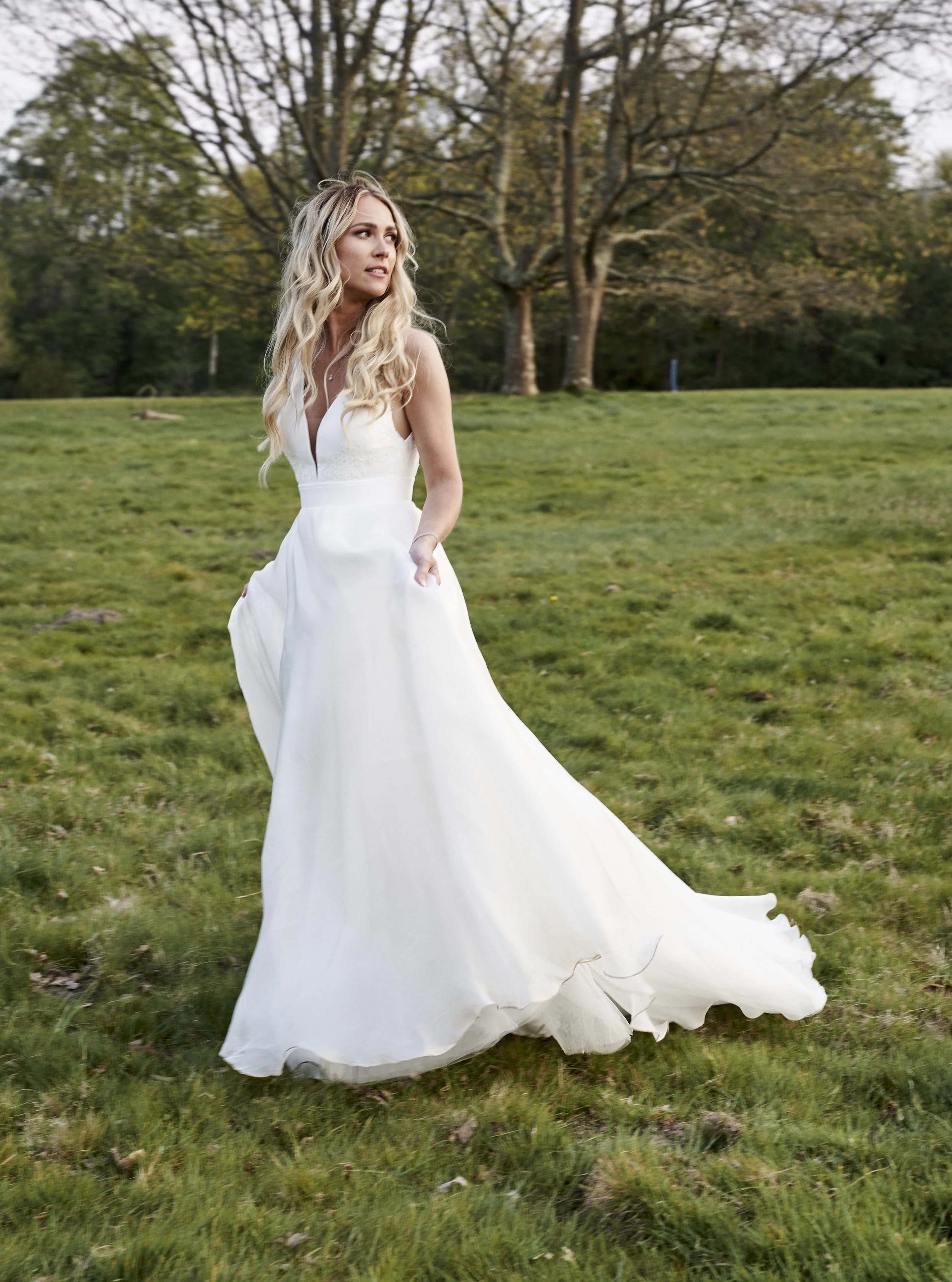 Woman in a white wedding dress standing on grass with trees in the background.