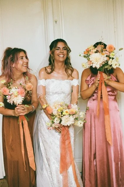Bride in a white wedding dress with two bridesmaids in earth-toned dresses holding flower bouquets, smiling and standing indoors.