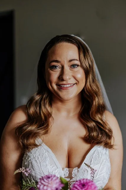 Bride smiling in a white lace wedding dress with a veil, holding a bouquet of flowers.