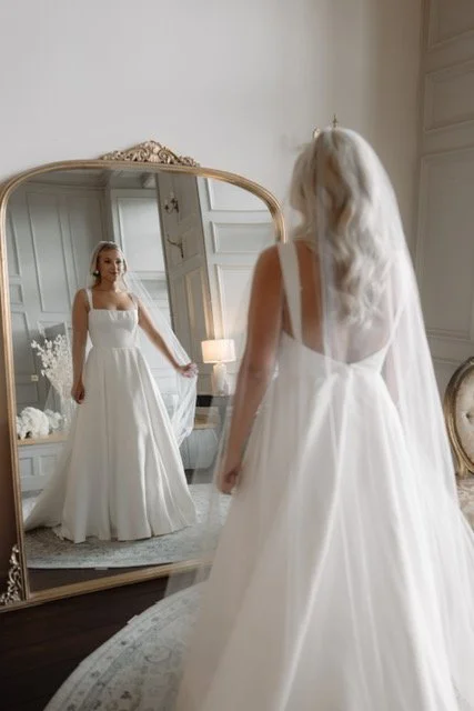 Bride in a white wedding dress standing in front of a large ornate mirror, looking at her reflection in an elegant room with paneled walls and soft lighting.