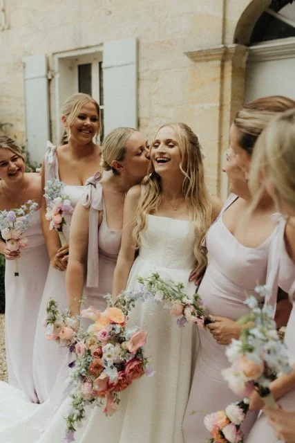 Bride and bridesmaids in light purple dresses holding bouquets and smiling outdoors.