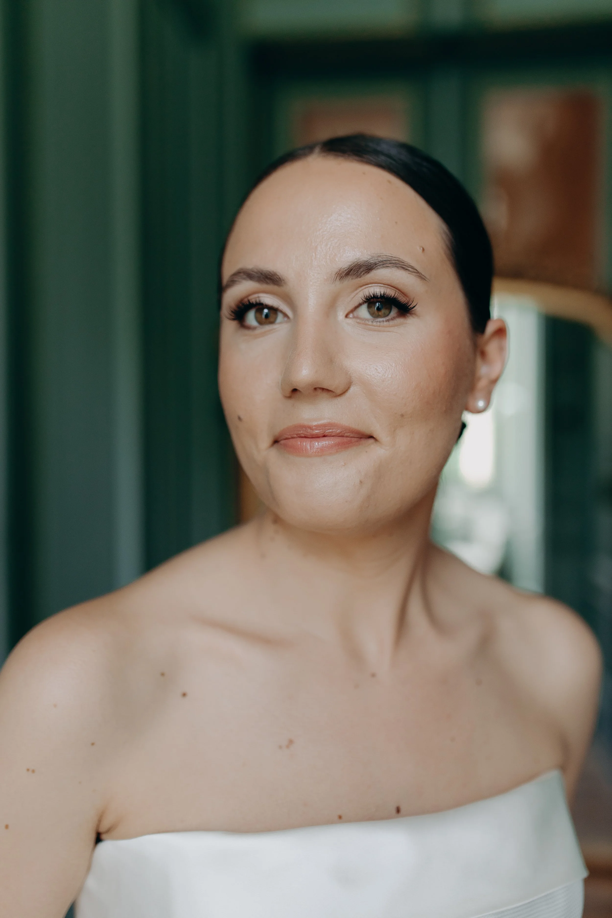 A woman with short dark hair and natural makeup wearing a strapless white dress, standing indoors.