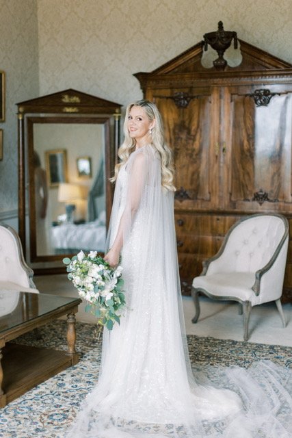 Bride in white dress holding bouquet, standing in vintage room with antique furniture and large mirror.