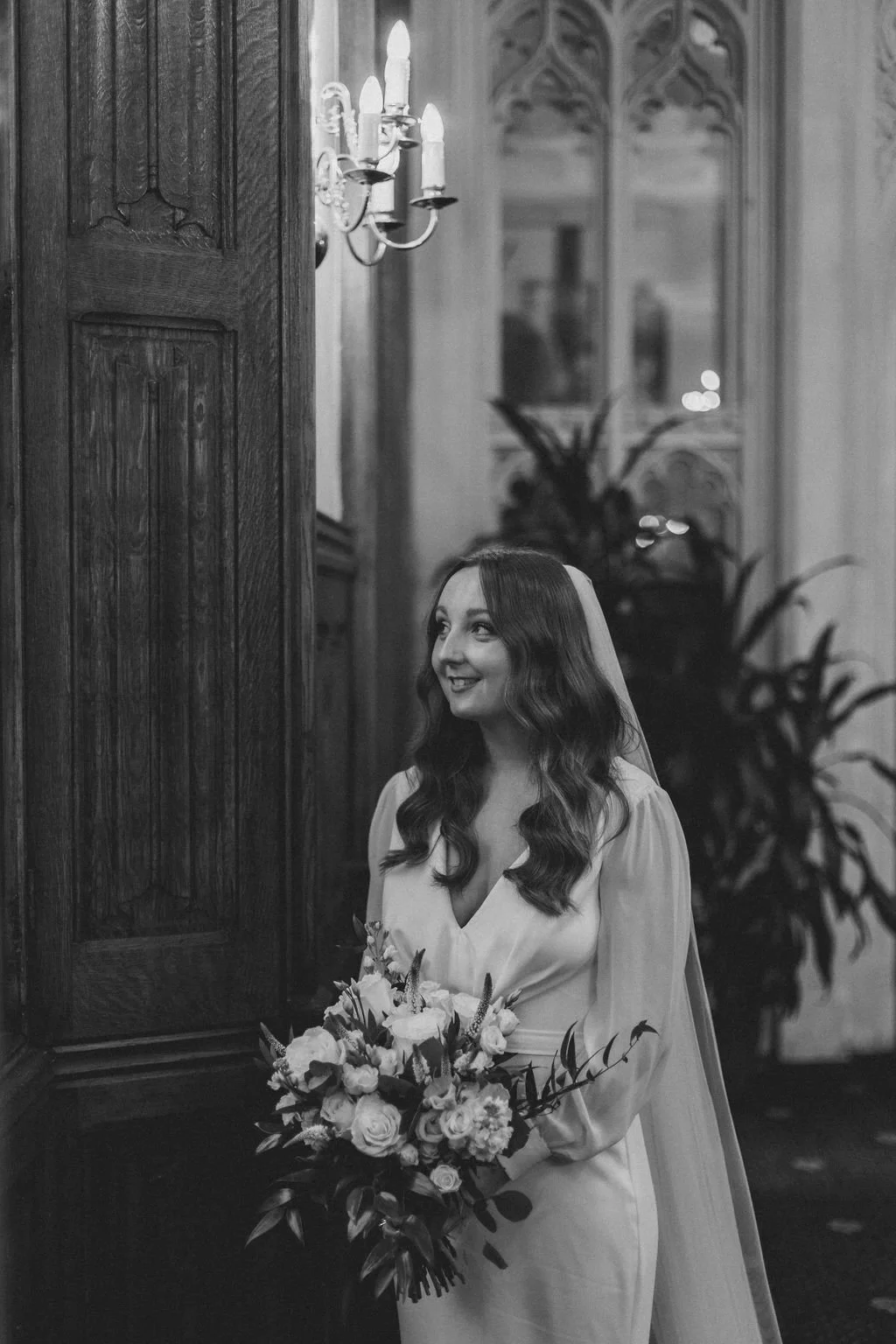 A black and white photo of a bride holding a bouquet, standing indoors by a wooden door, with a sconce and ornate window in the background.