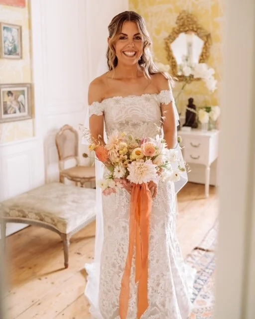 Bride in white lace wedding gown holding a bouquet with pink and orange flowers in vintage-style room.