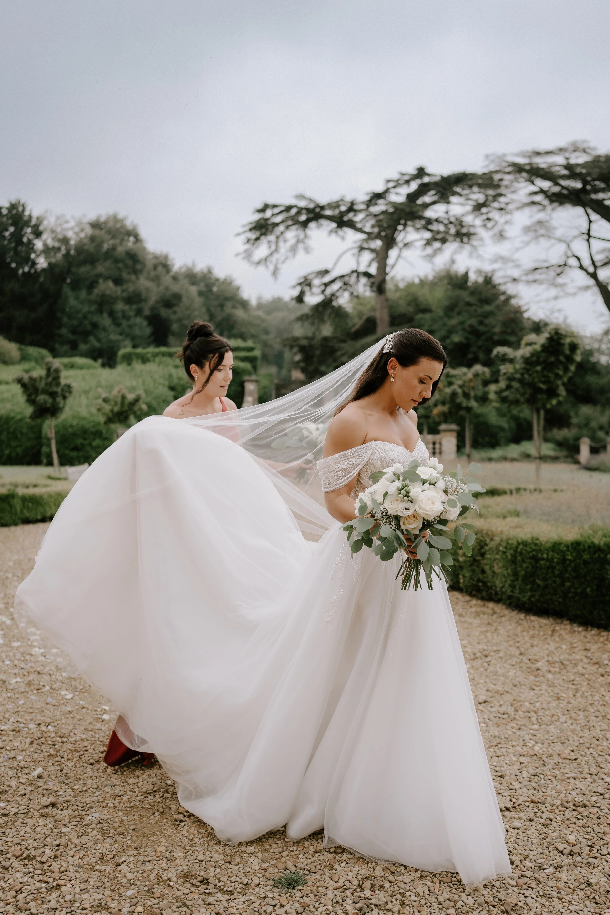 Bride in a white wedding dress holding a bouquet, with a bridesmaid adjusting the dress train, set in an outdoor garden setting.