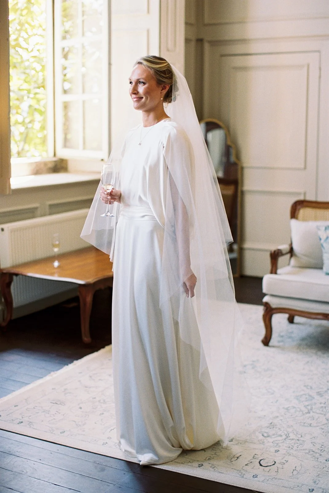 Bride wearing a white wedding dress with a long veil, holding a champagne glass, standing in a bright room with a window and antique furniture.