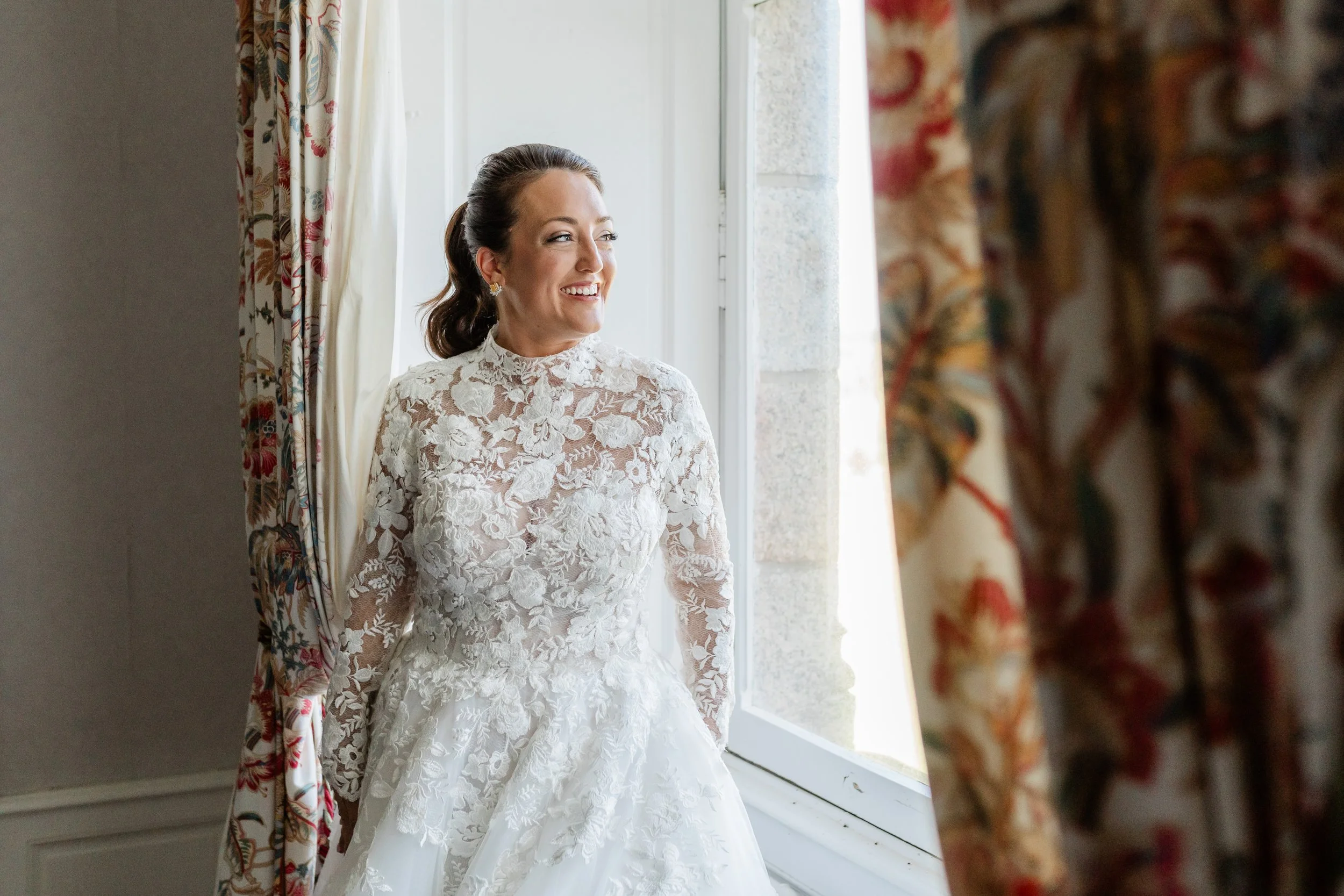 Bride in lace wedding dress smiling near window with floral curtains