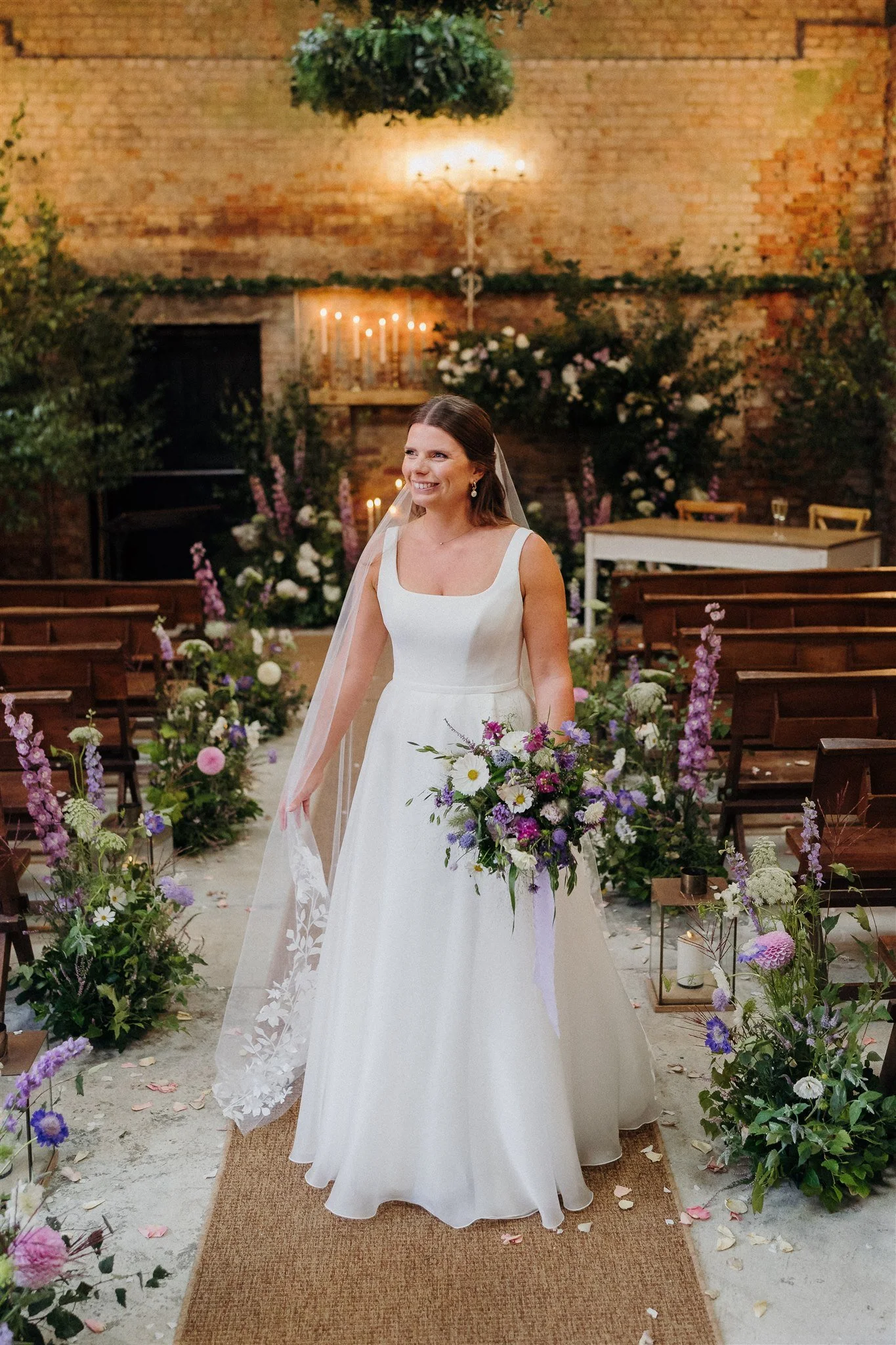 Bride standing in wedding aisle with floral decorations, holding a bouquet, surrounded by wooden pews and greenery, in an indoor venue with a brick wall backdrop.