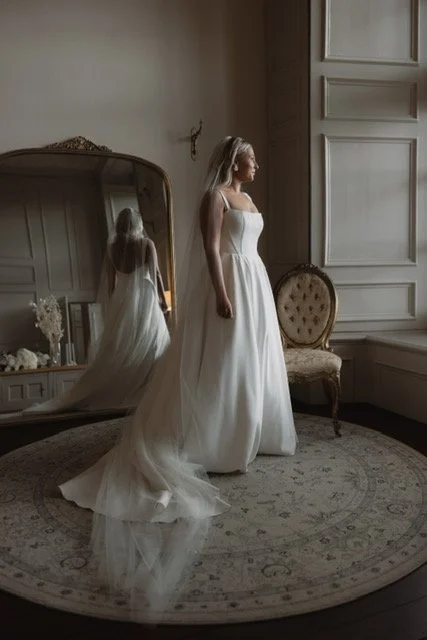A bride in a white wedding gown stands in a stylish room with a large mirror, vintage chair, and ornate rug.