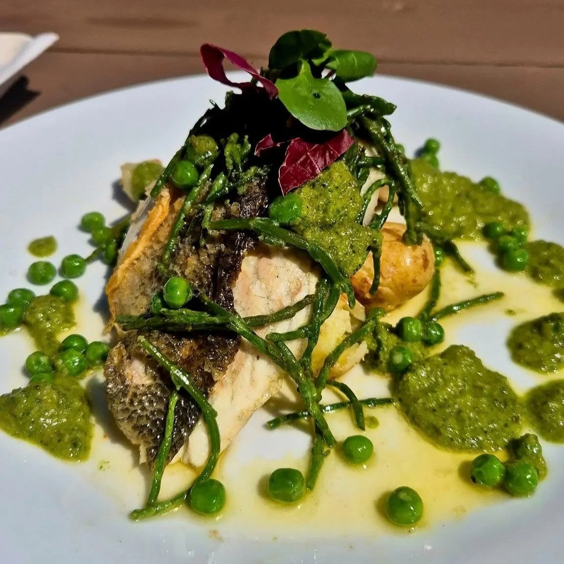 Plate with cooked fish fillet topped with green herbs, peas, and green sauce, garnished with microgreens.