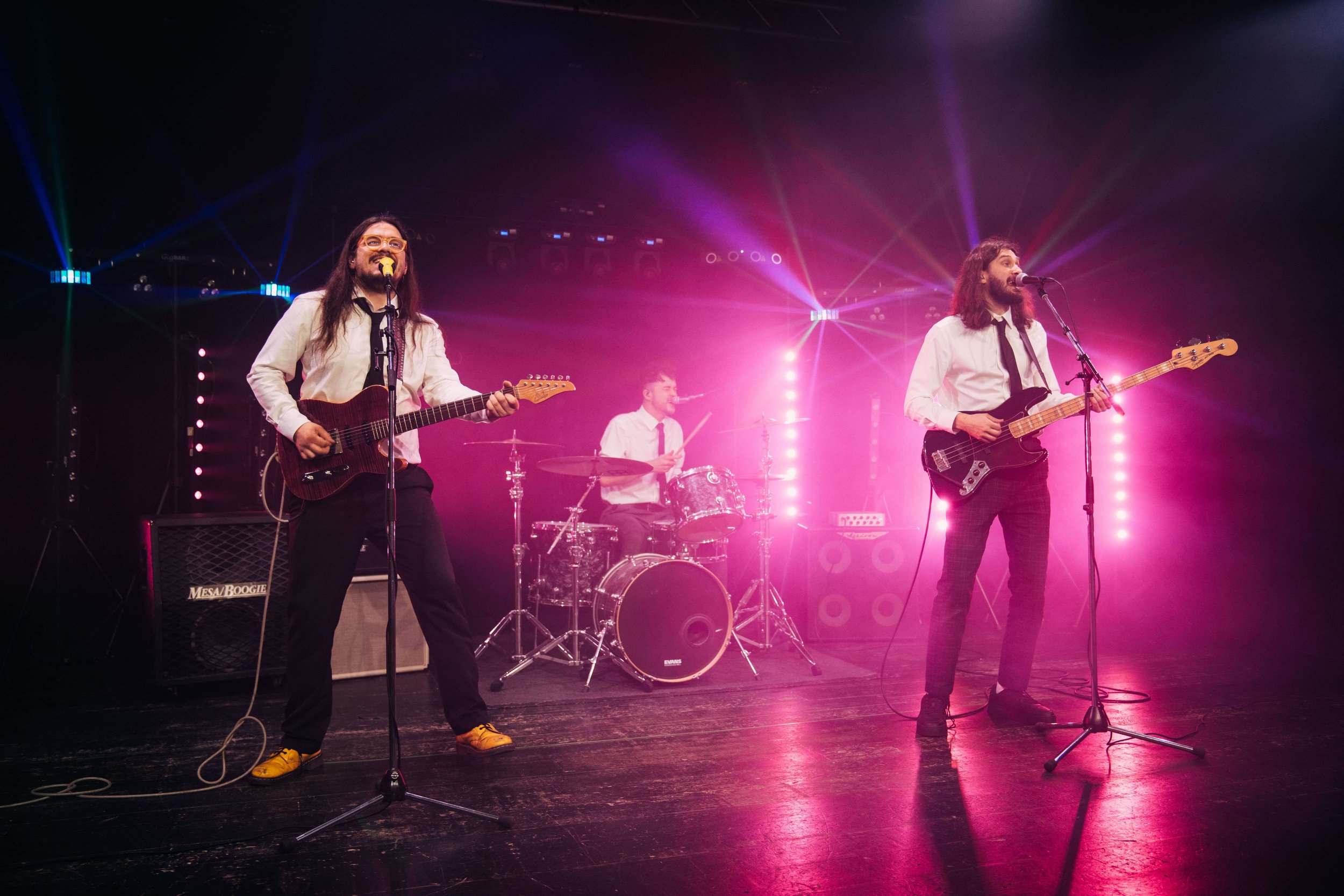 A band performing on stage with two guitarists and a drummer, colorful stage lights, and fog effects.