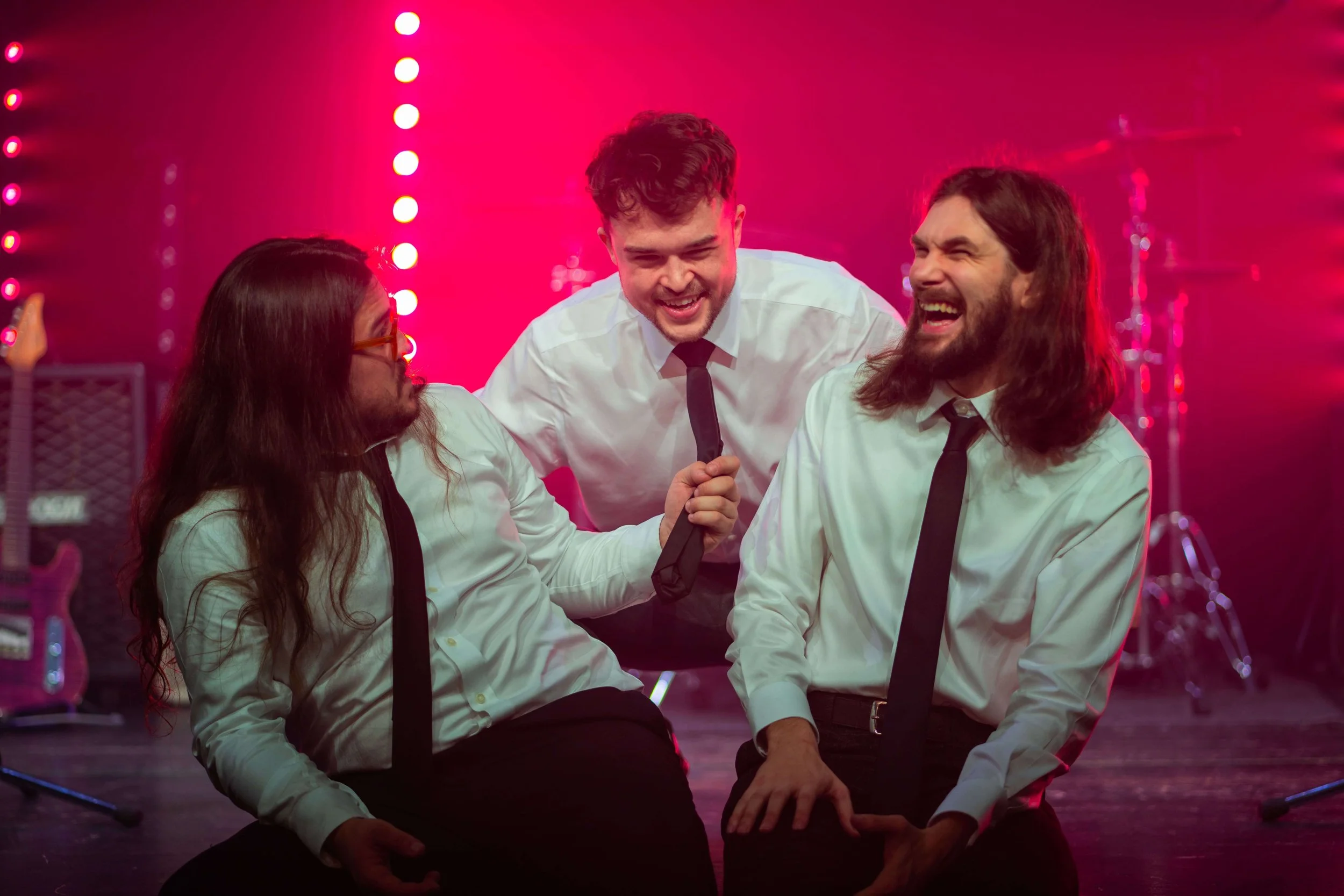Three men in white shirts and black ties, laughing and talking on stage with pink and purple lighting, musical instruments in the background.