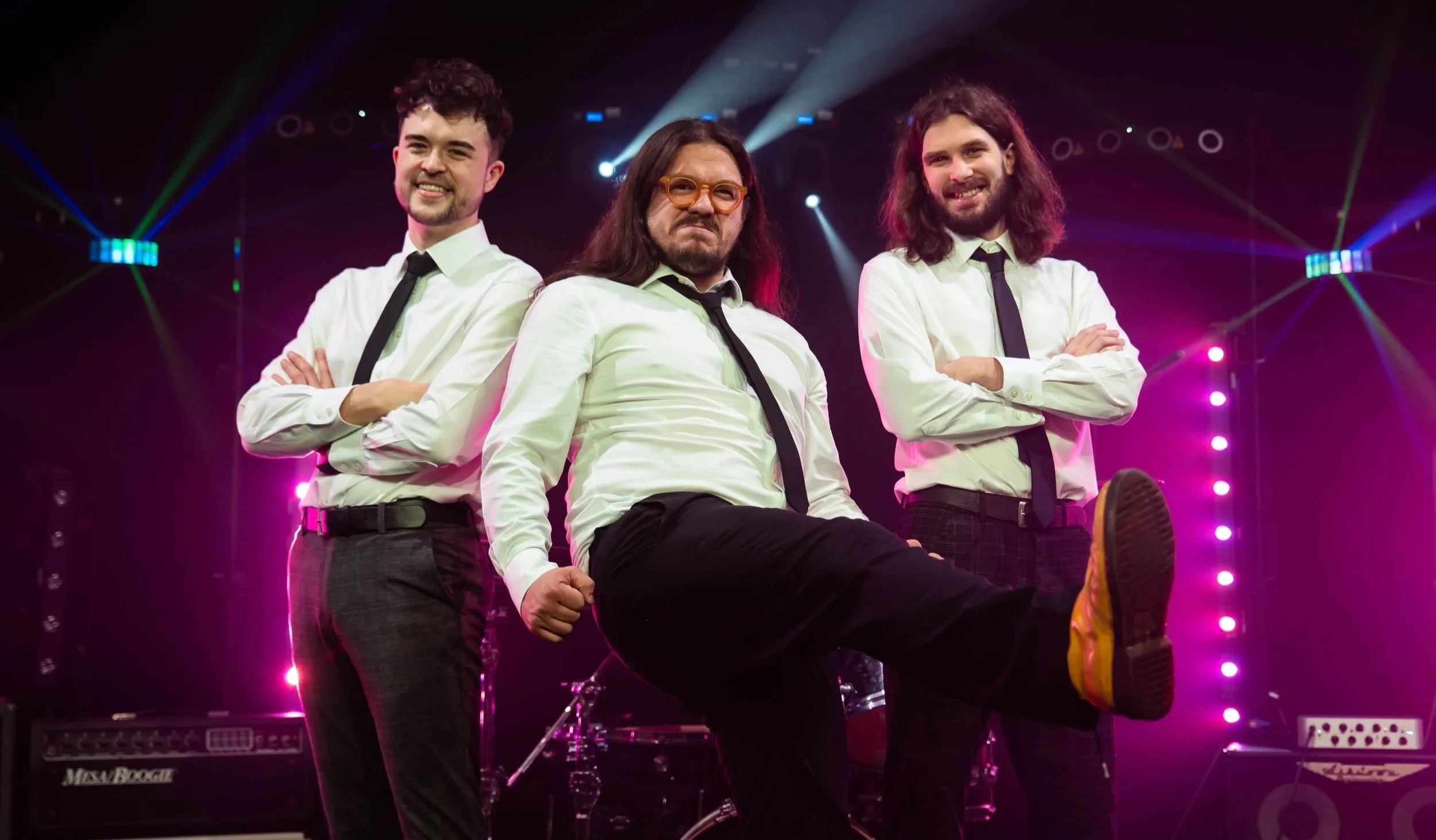 Three men in white shirts and black ties posing on stage with musical equipment and purple lights in the background, one man standing with arms crossed and smiling, another man with long hair and beard also smiling, and a third man with glasses and long hair with an intense expression, one man has his leg raised.