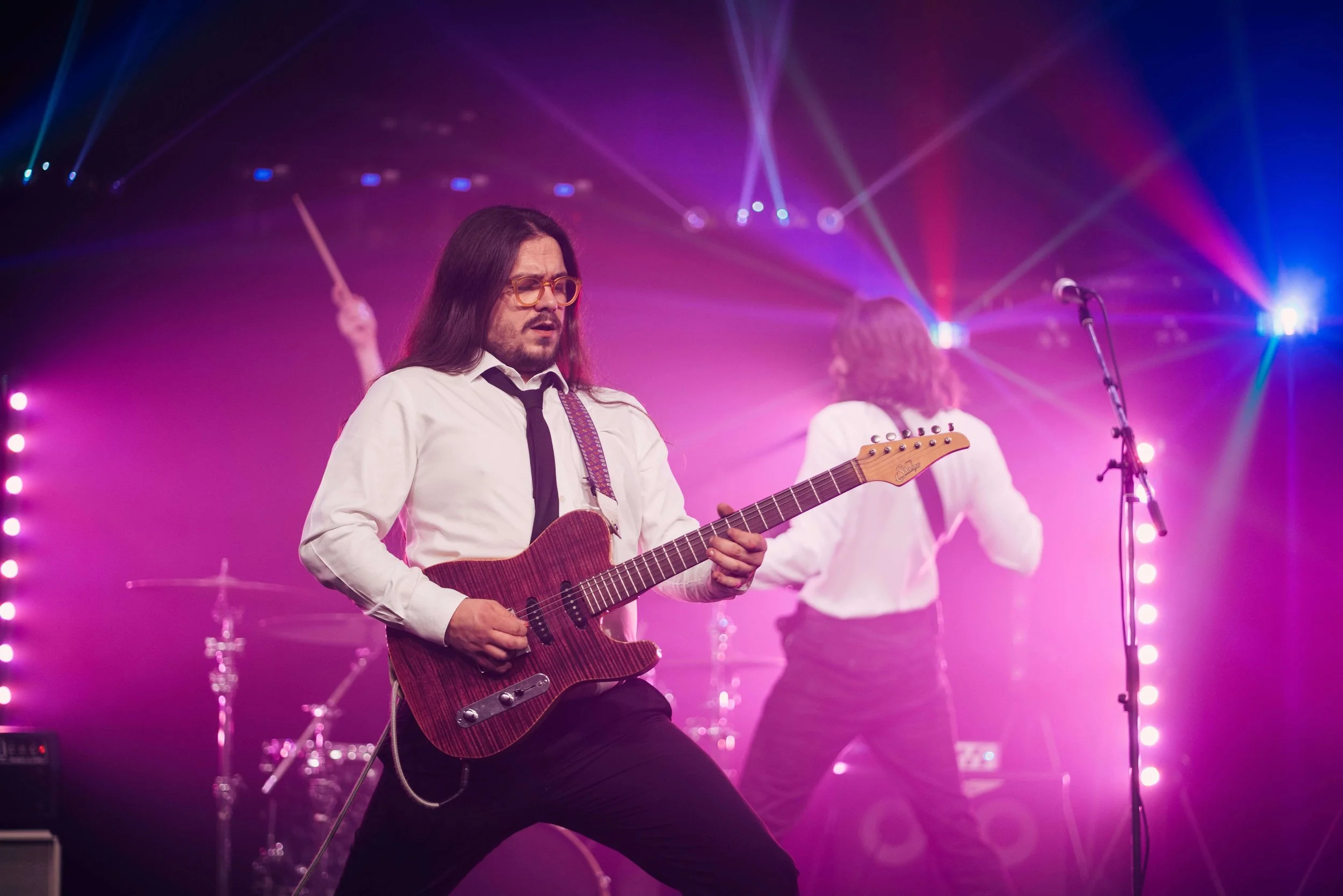 Musician playing electric guitar on stage with colorful lighting effects and fog, with another musician in the background.