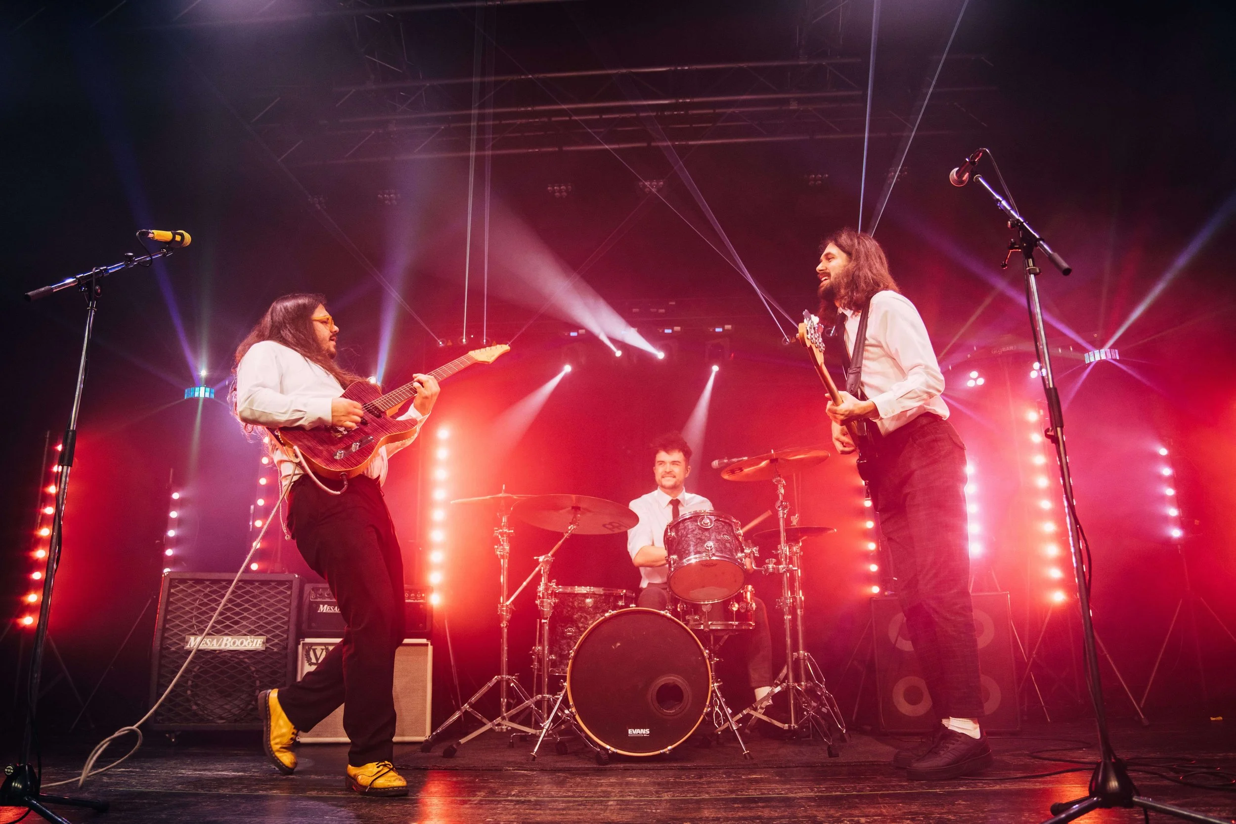 Three musicians performing on stage with colorful lighting and microphones, one playing guitar, one on drums, and one playing bass guitar.