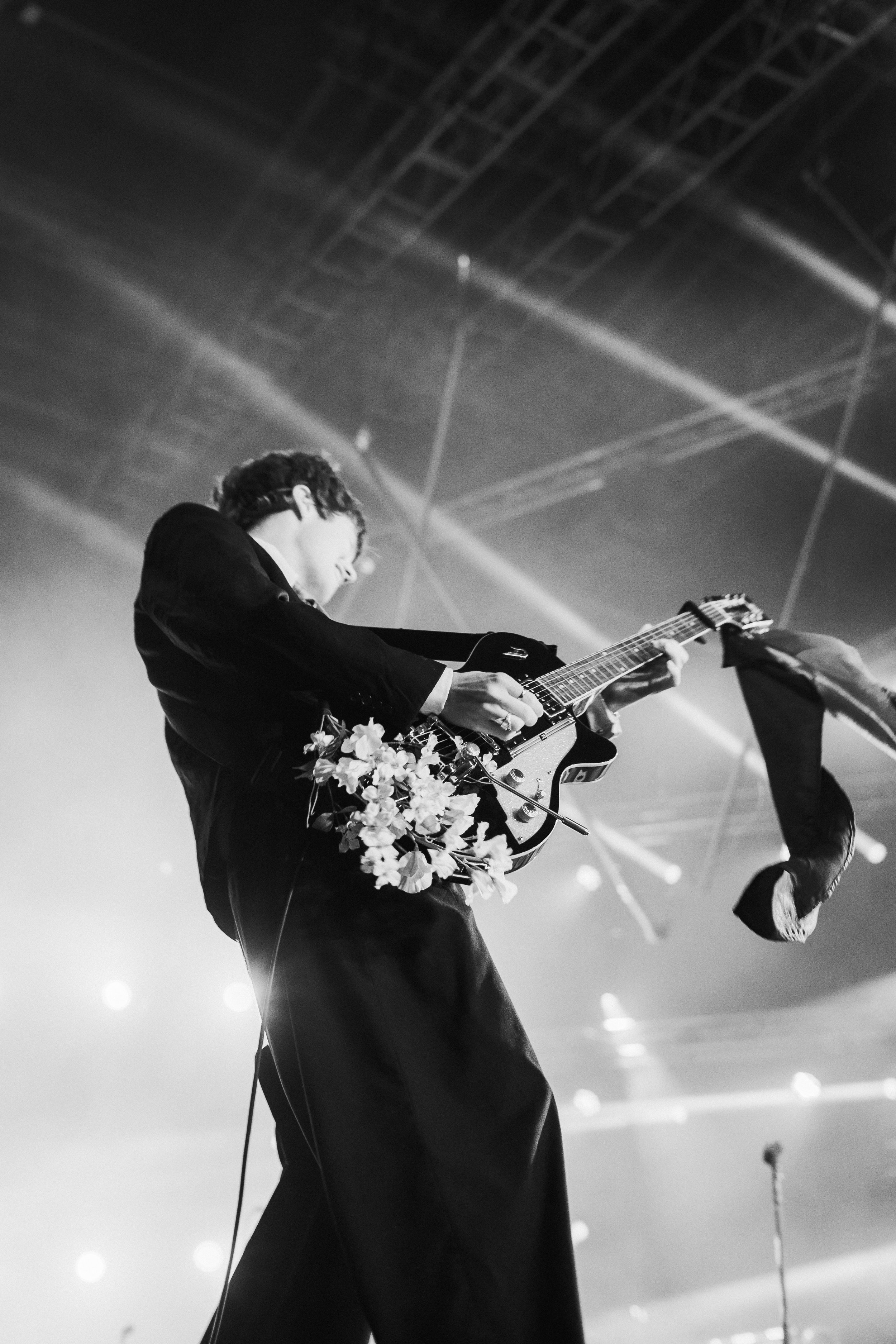A musician in a tuxedo performing on stage with an electric guitar decorated with flowers, captured in black and white from a low angle.