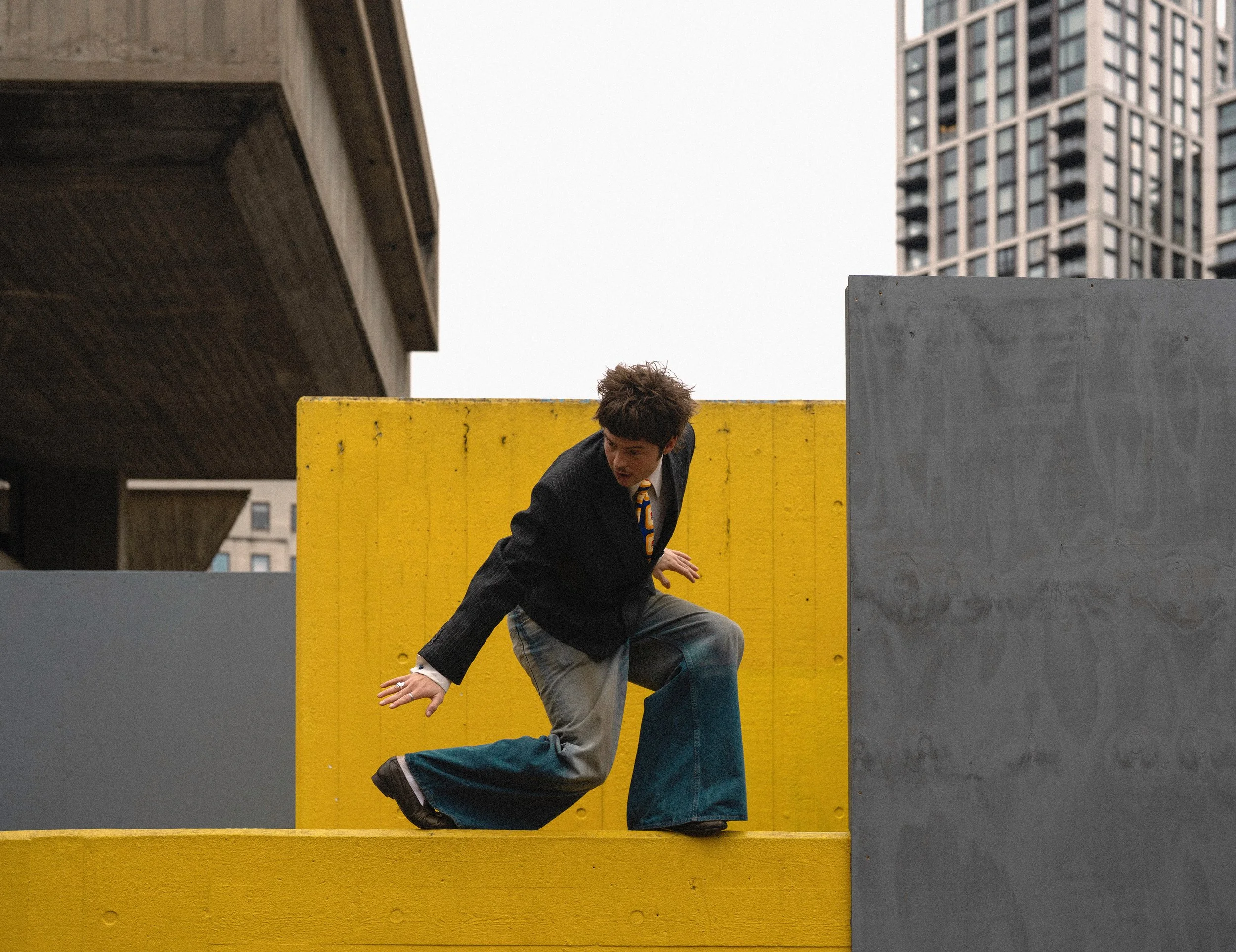 A person in a black blazer, white shirt, and wide-leg jeans crouches on a yellow structure in an urban setting. The person has short, curly hair and appears to be looking to the side. There are tall buildings in the background, and the sky is overcast.