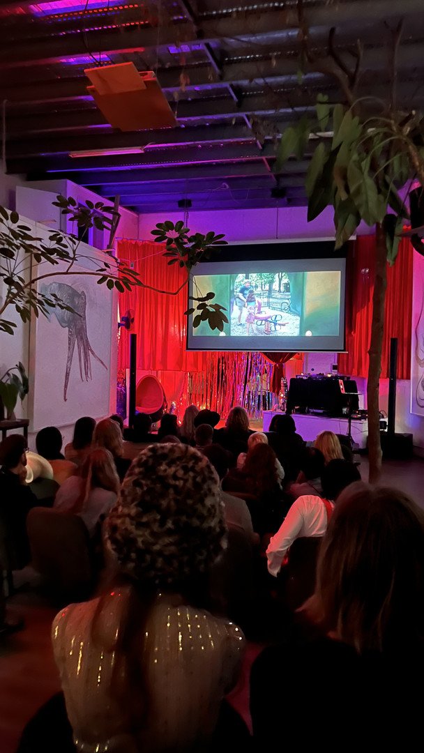 Indoor event with audience watching a projected film; colorful lighting and decorations, including plants and a painted wall.