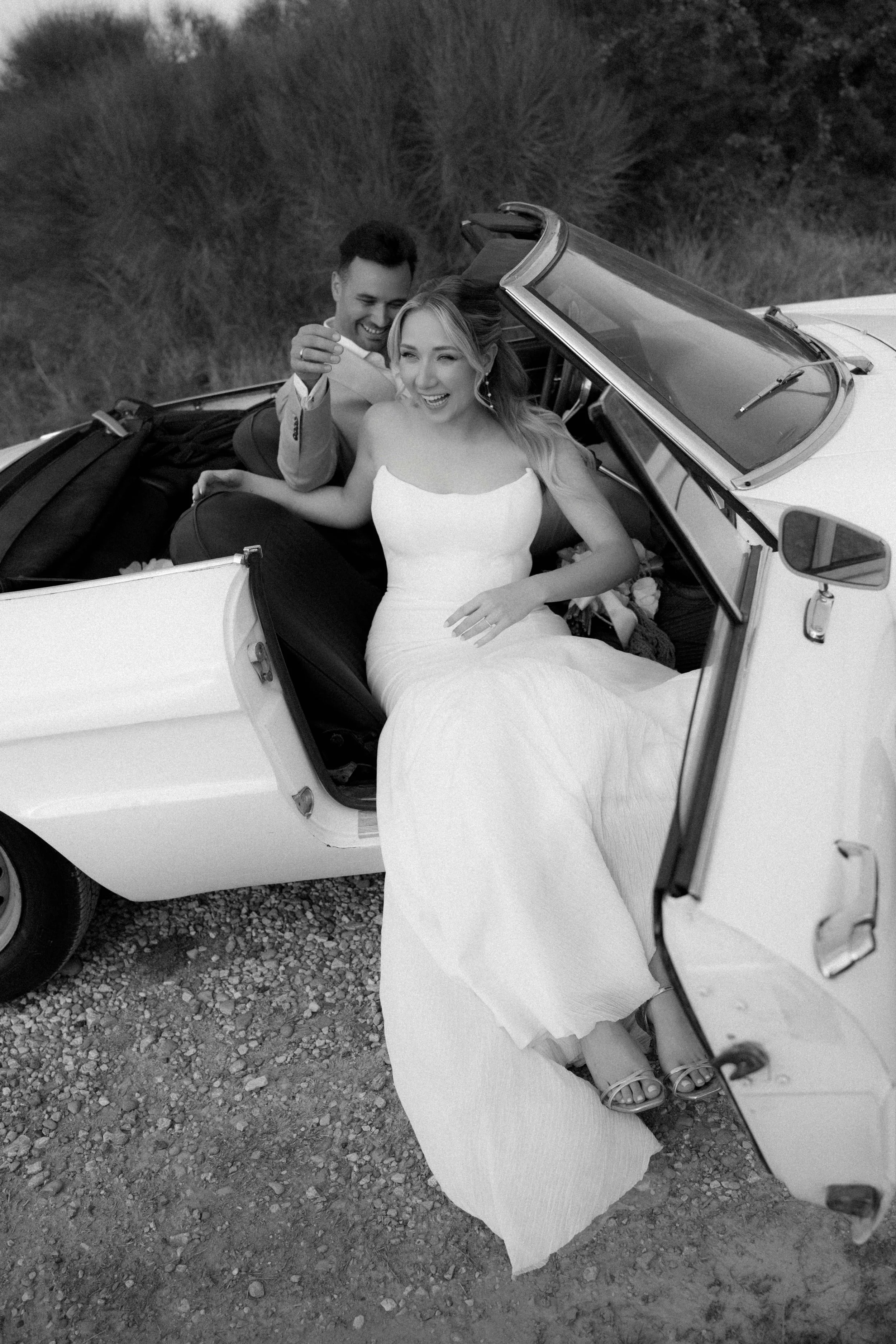 Ein lächelndes Brautpaar sitzt in einem Cabrio. Die Braut trägt ein weißes Hochzeitskleid und Sandalen, der Bräutigam trägt einen grauen Anzug. Das Auto ist auf einer graveligen Straße.
