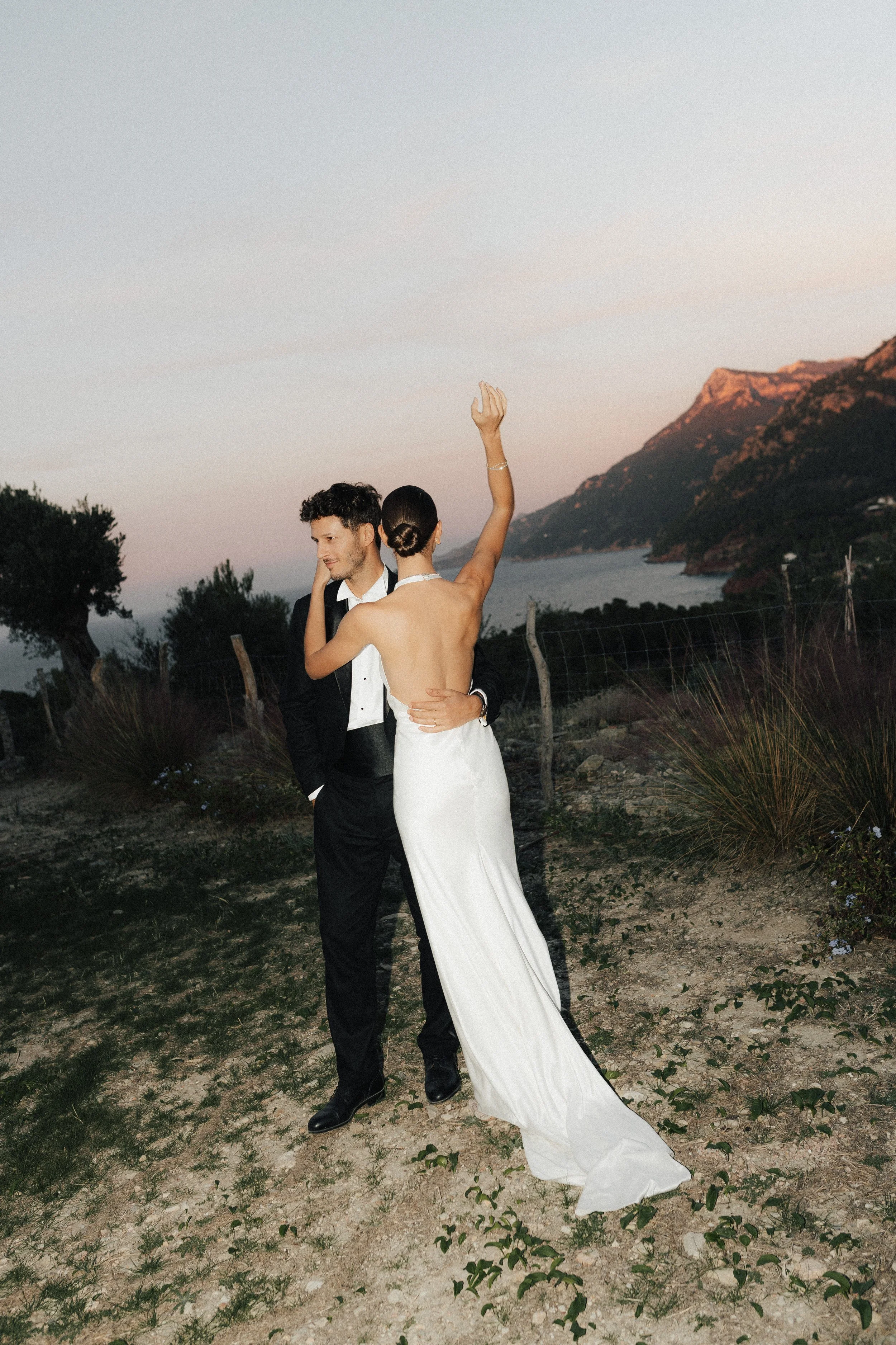Ein Paar bei einer romantischen Hochzeit im Freien bei Sonnenuntergang, im Hintergrund Berge und Wasser. Die Frau trägt ein langes weißes Brautkleid, der Mann einen schwarzen Anzug, sie halten sich liebevoll umarmend.
