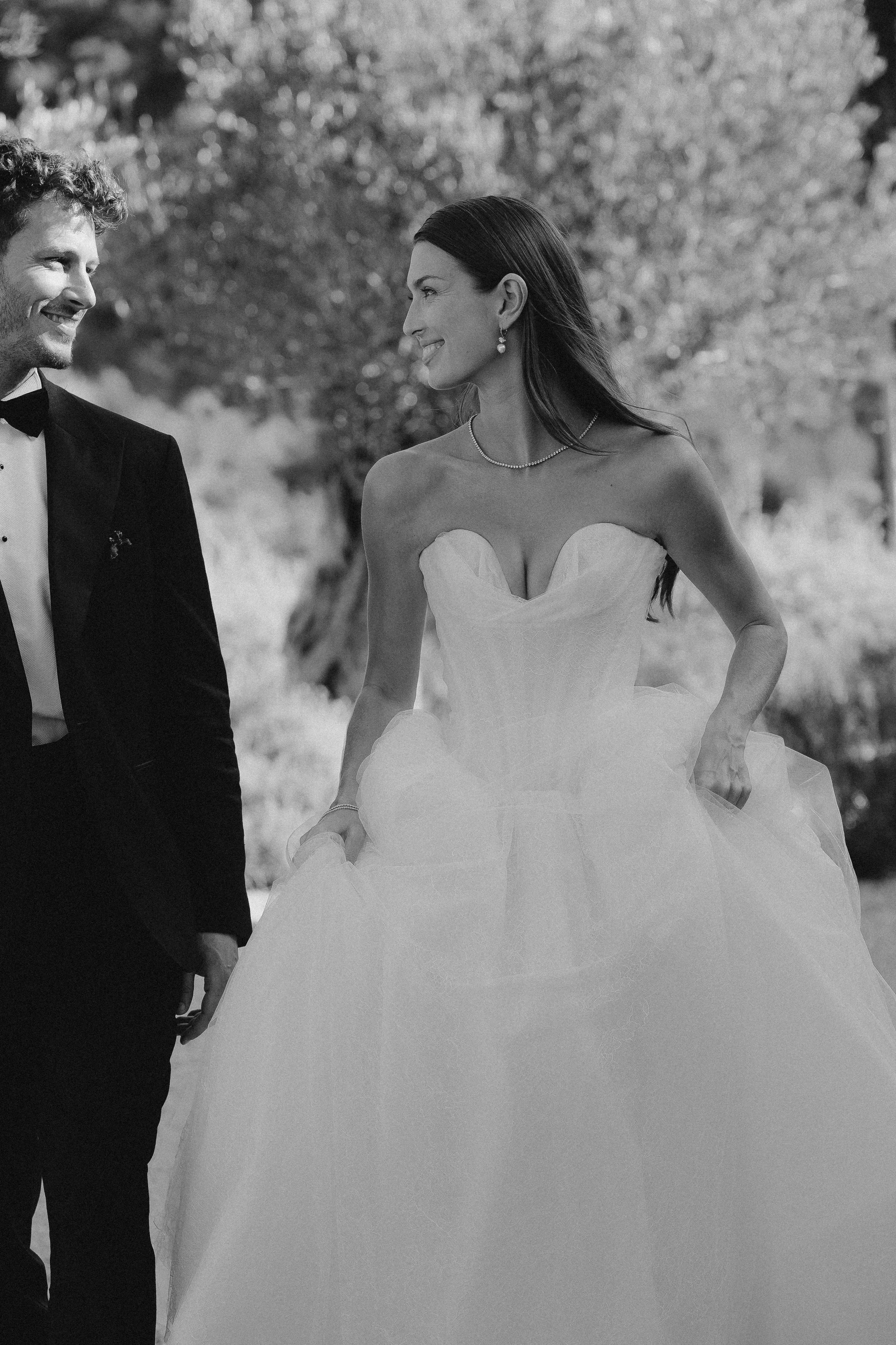 Ein Brautpaar bei ihrer Hochzeit, die Frau trägt ein Hochzeitskleid und der Mann einen Smoking, beide lachen sich an, im Freien bei Bäumen.