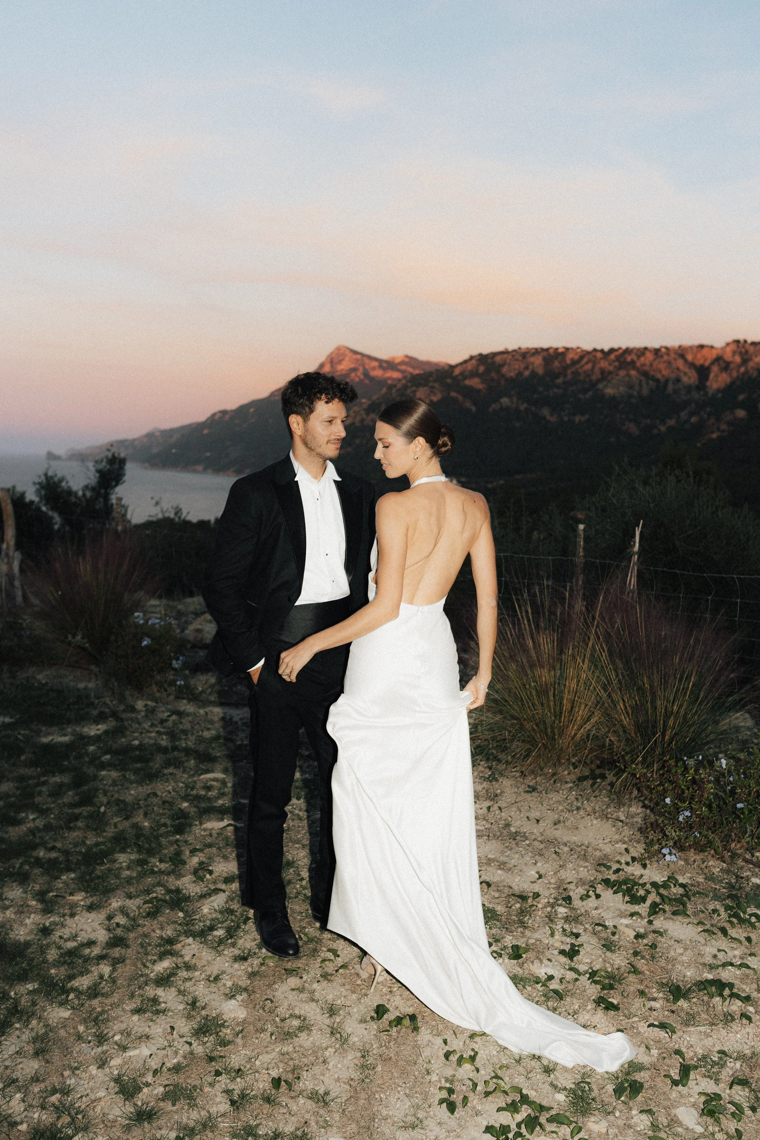 Ein Mann und eine Frau in Hochzeitskleidung stehen sich an einem Abend in einer ländlichen Gegend mit Bergen und Wasser im Hintergrund gegenüber.