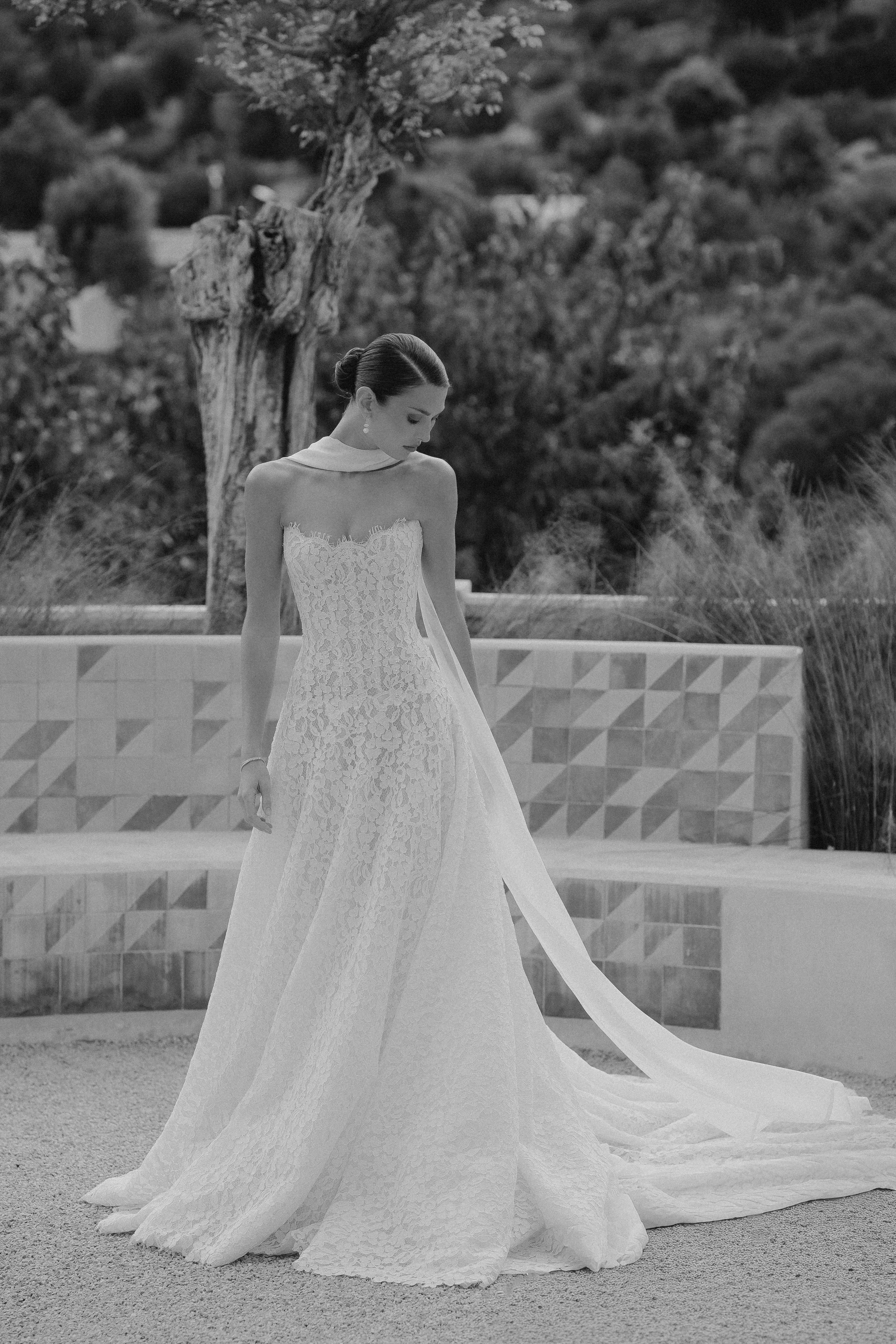 Eine Frau in einem eleganten, weißen Hochzeitskleid steht im Freien vor einem Baum, im Schwarz-Weiß-Stil.