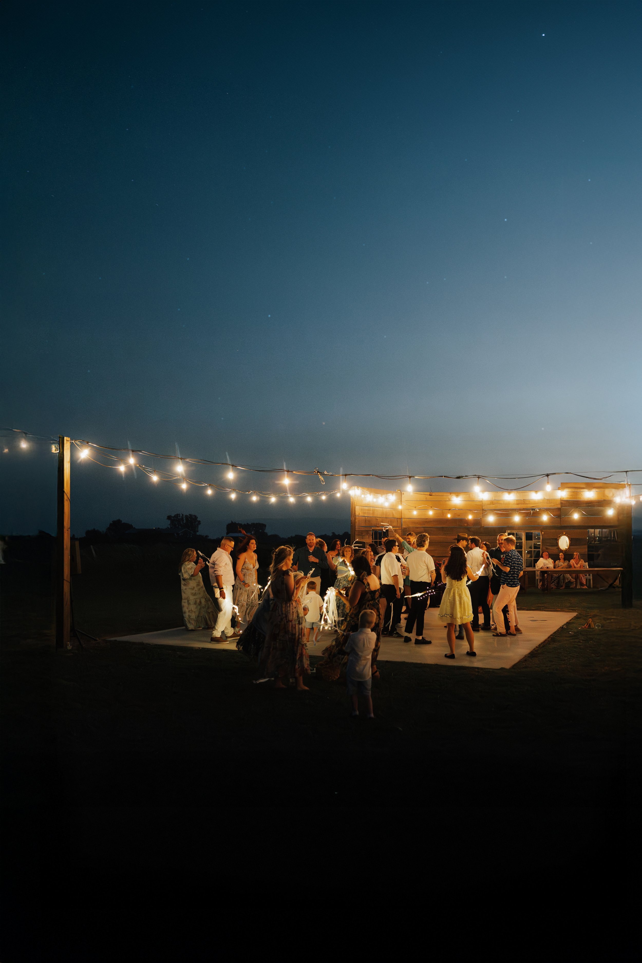 Menschen feiern bei einer nächtlichen Veranstaltung im Freien unter Lichterketten auf einer Plattform, mit einem dunklen Himmel im Hintergrund.