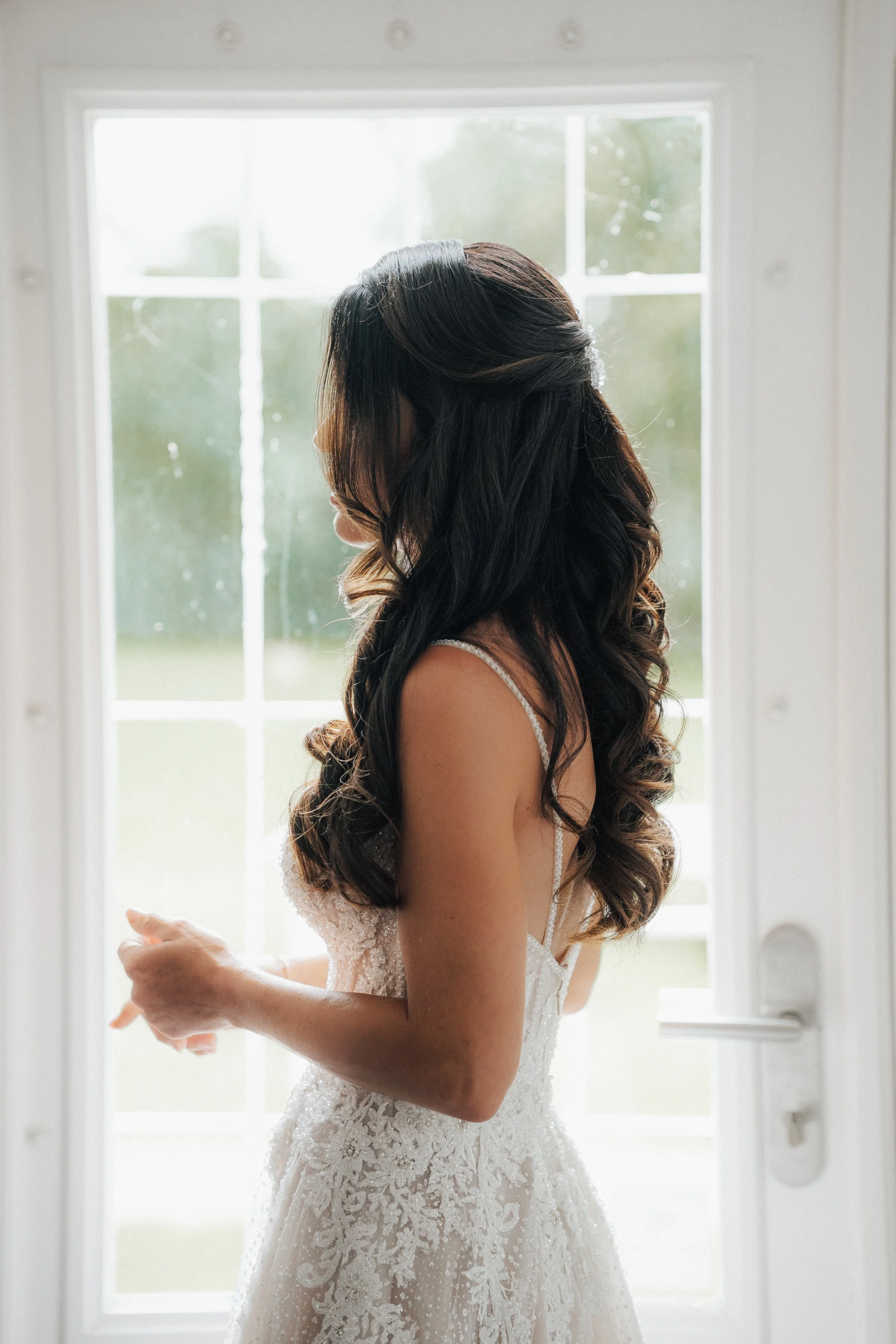 Frau im weißen Brautkleid mit schwarzen, lockigen Haaren steht seitlich vor einer weißen Tür mit Fenstern, im Hintergrund ist eine Grünfläche sichtbar.