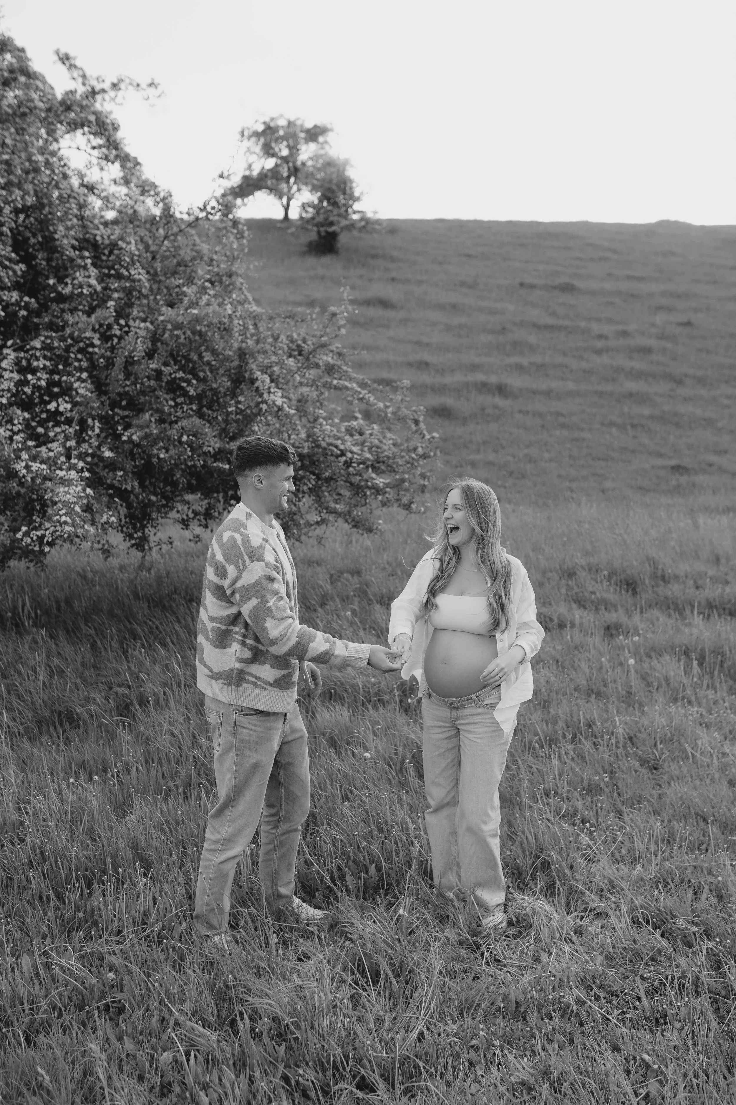 Ein Paar in einem Feld, die schwangere Frau hält die Hand ihres Partners, beide lachen und sind glücklich.