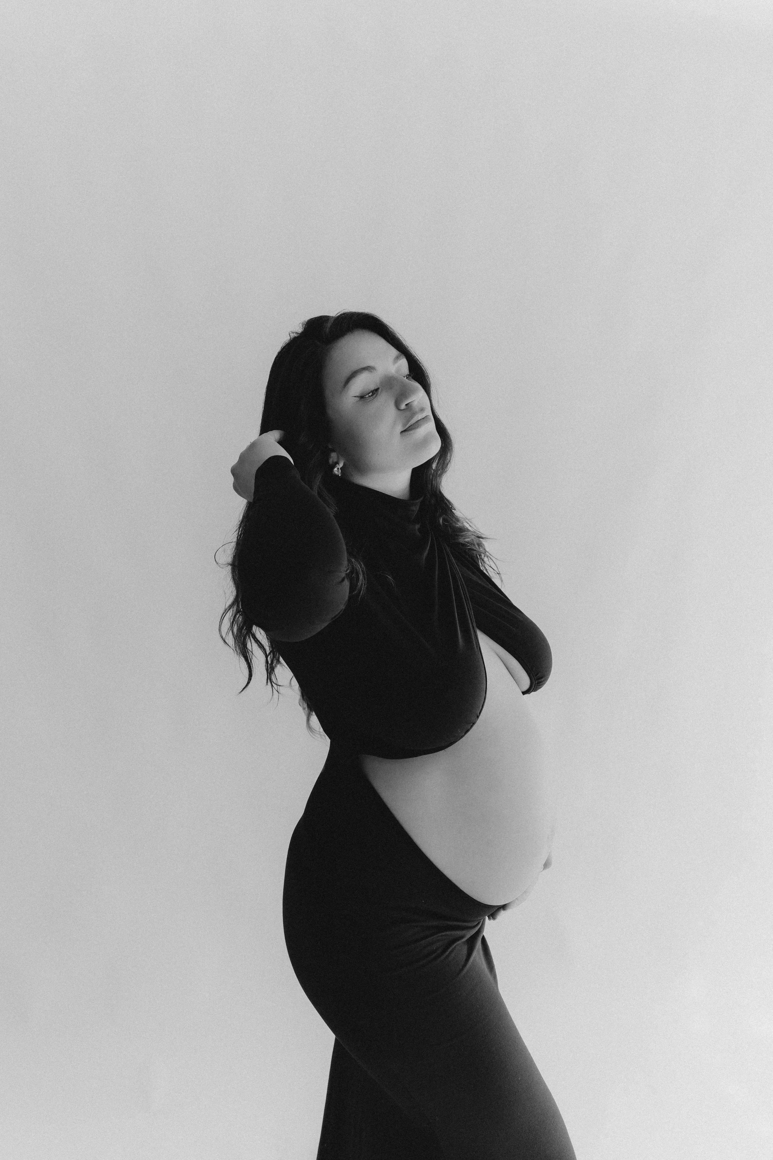 Schwangere Frau in schwarzen Kleid vor weißem Hintergrund, blickt seitlich, hat dunkle, lange Haare, Hand im Haar, entspannt.