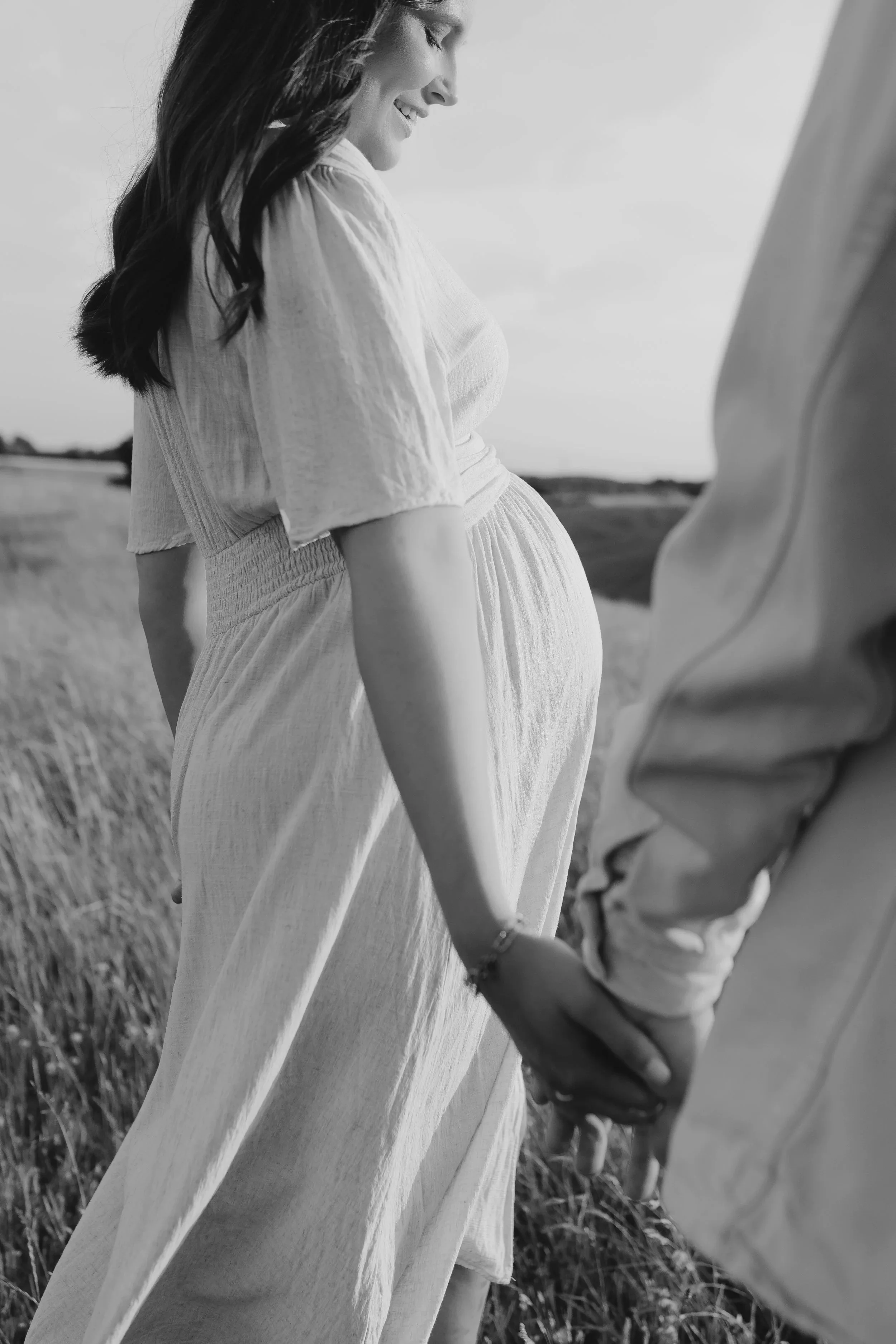 Schwangere Frau hält die Hand ihres Partners in einem Feld im Freien, beide lächeln, schwarz-weiß Fotografie.