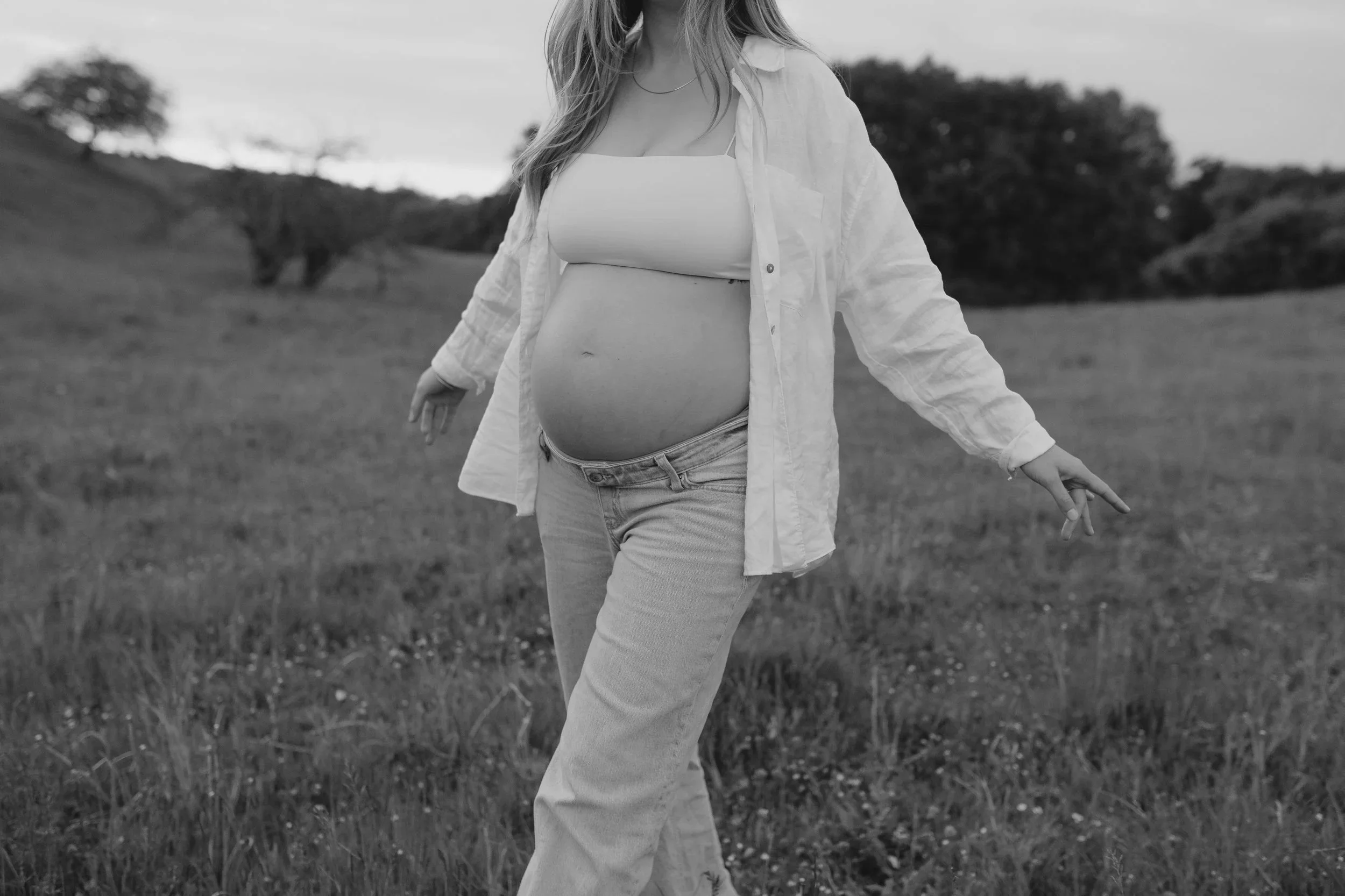 Eine schwangere Frau in einem Feld, sie trägt eine helle Jacke, ein top und Hose, in Schwarz-Weiß aufgenommen.