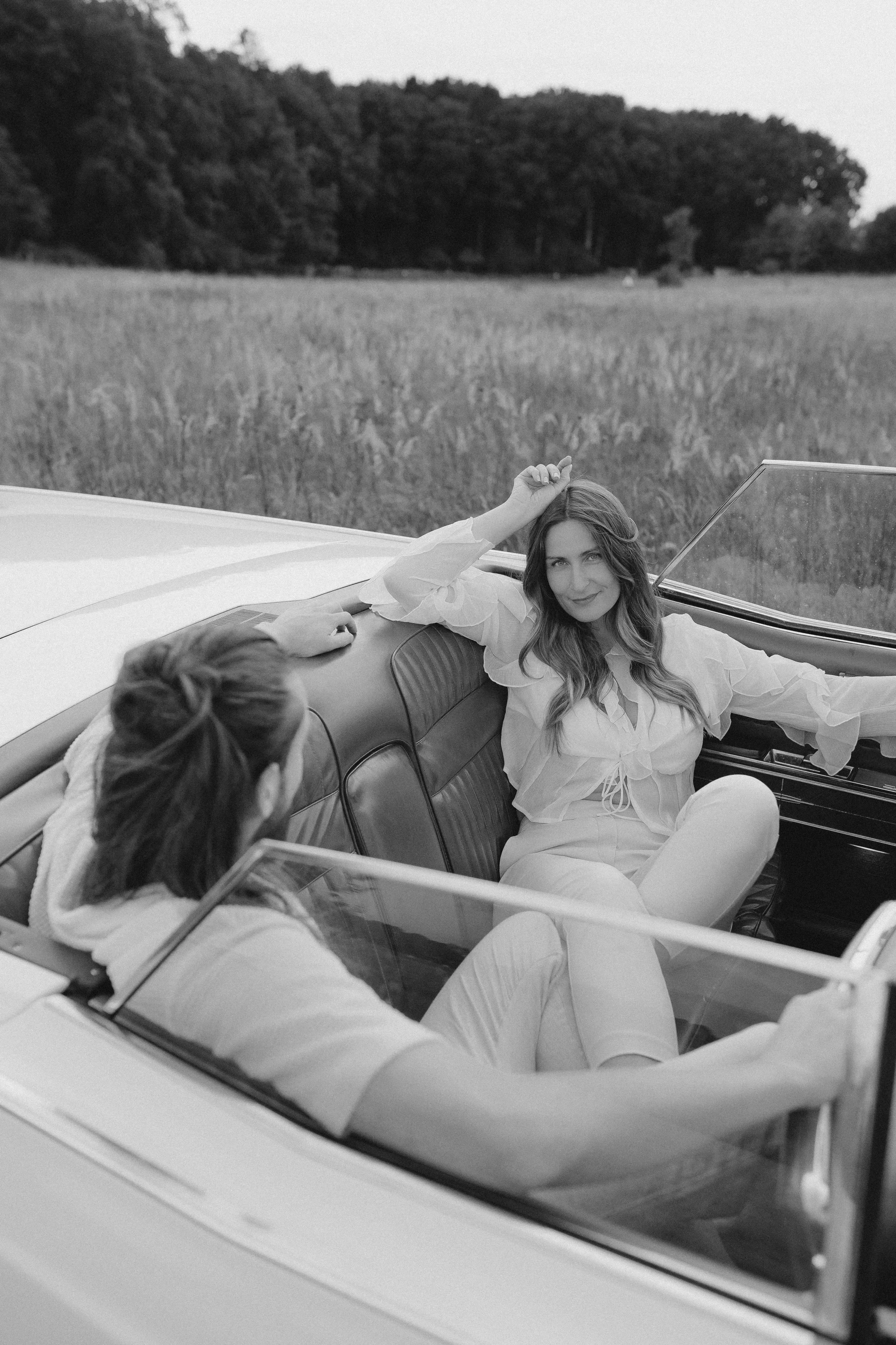 Zwei Frauen sitzen in einem klassischen Auto auf dem Land, eine von ihnen lächelt in die Kamera, die andere blickt weg. Die Szene ist schwarz-weiß.