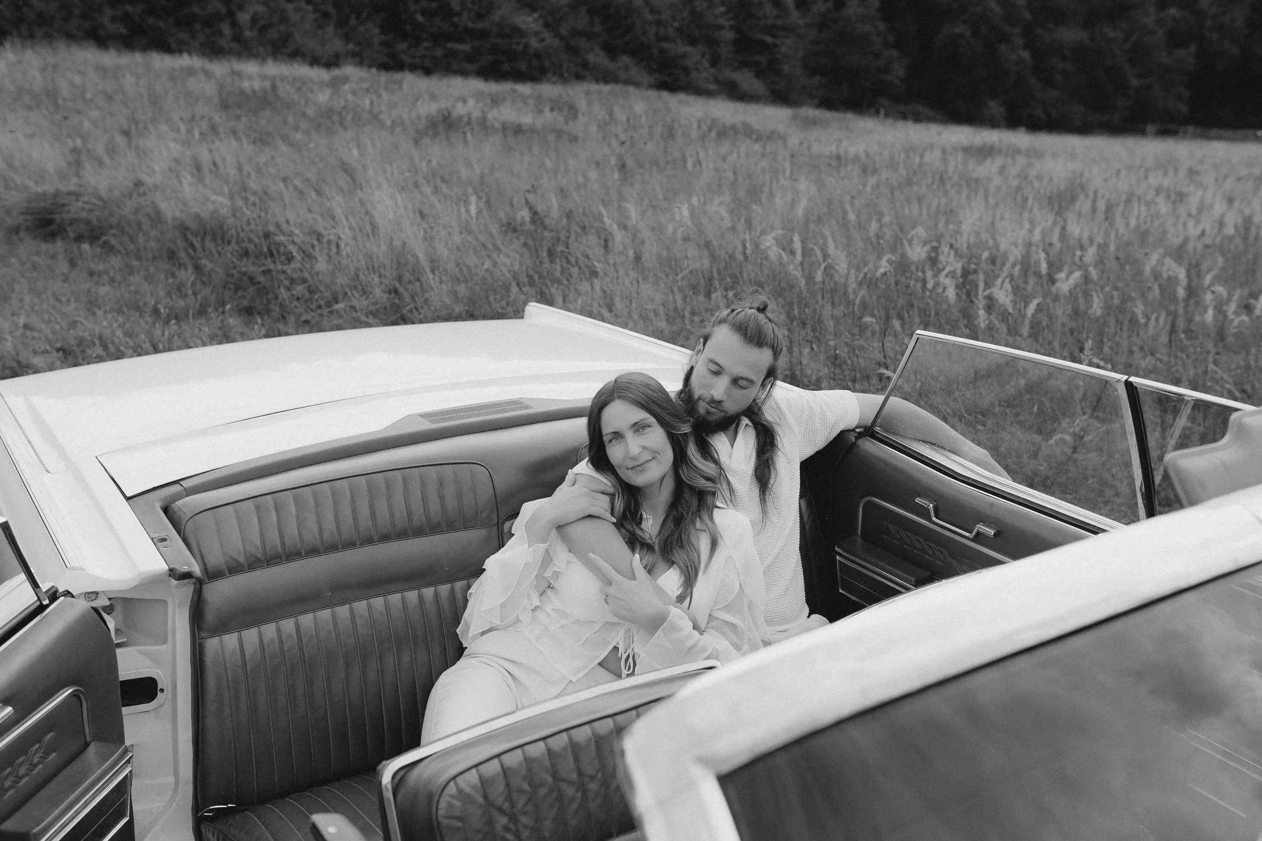 Ein Mann und eine Frau sitzen in einem offenen Oldtimer in einem Feld, umgeben von Natur, schwarz-weiß aufgenommen.