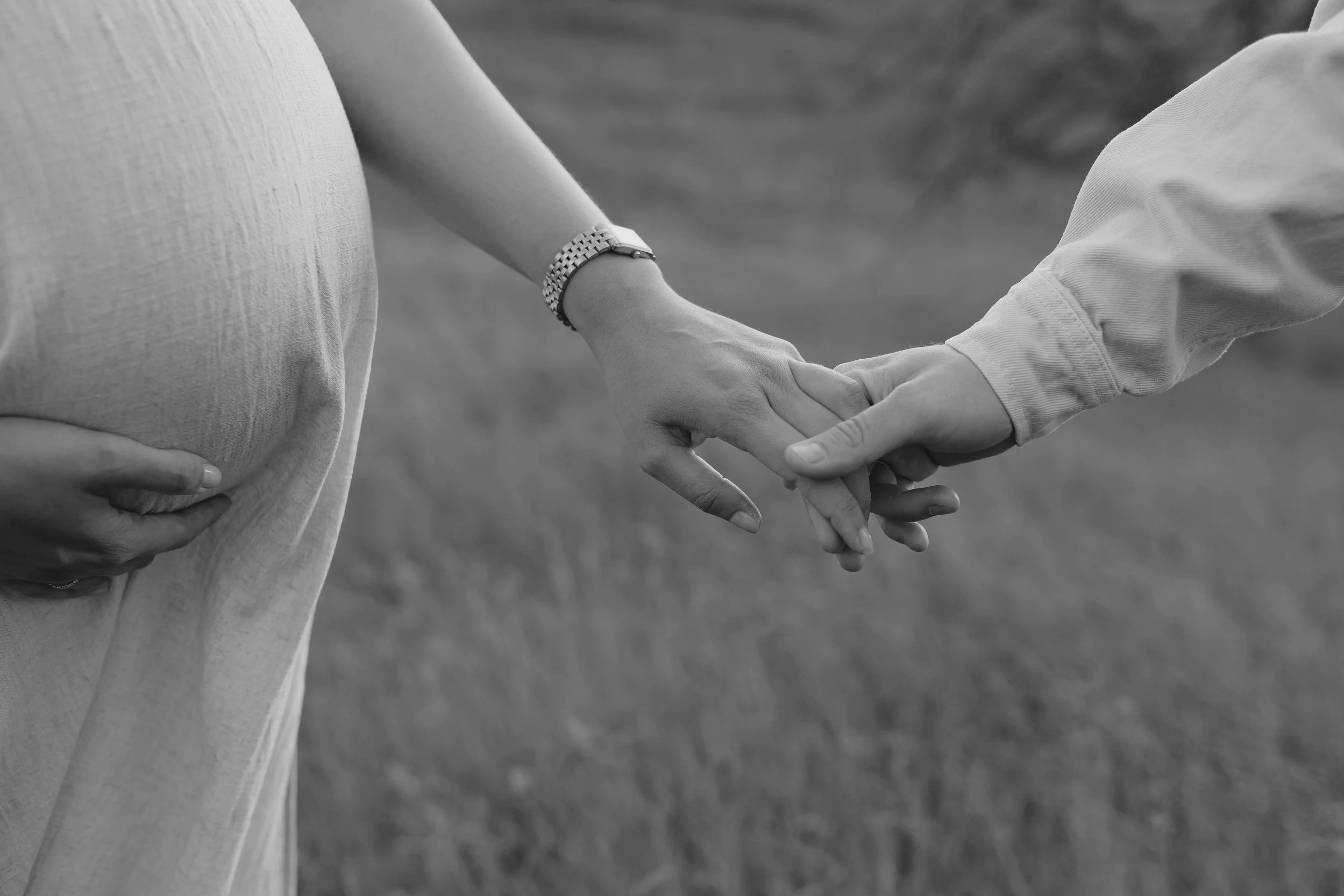Zwei Menschen halten sich an der Hand und eine Person berührt ihren Bauch, vermutlich eine Schwangerschaft, im Hintergrund ist eine Wiese oder ein Feld, schwarz-weiß Foto.
