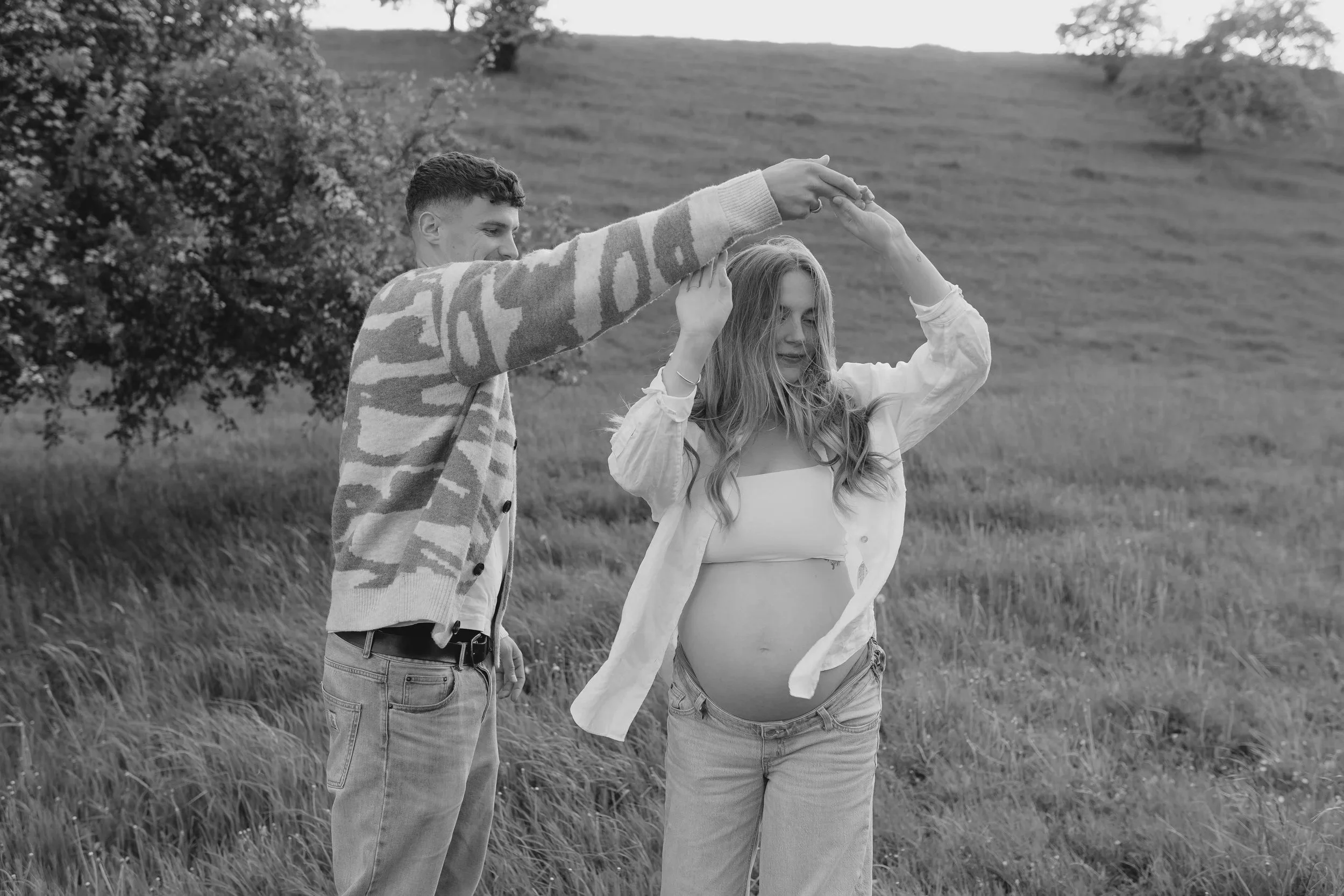 Ein Mann und eine schwangere Frau stehen in einer Wiese. Der Mann formt einen Kreis mit den Armen über den Kopf der Frau, die mit geschlossenen Augen lächelt.