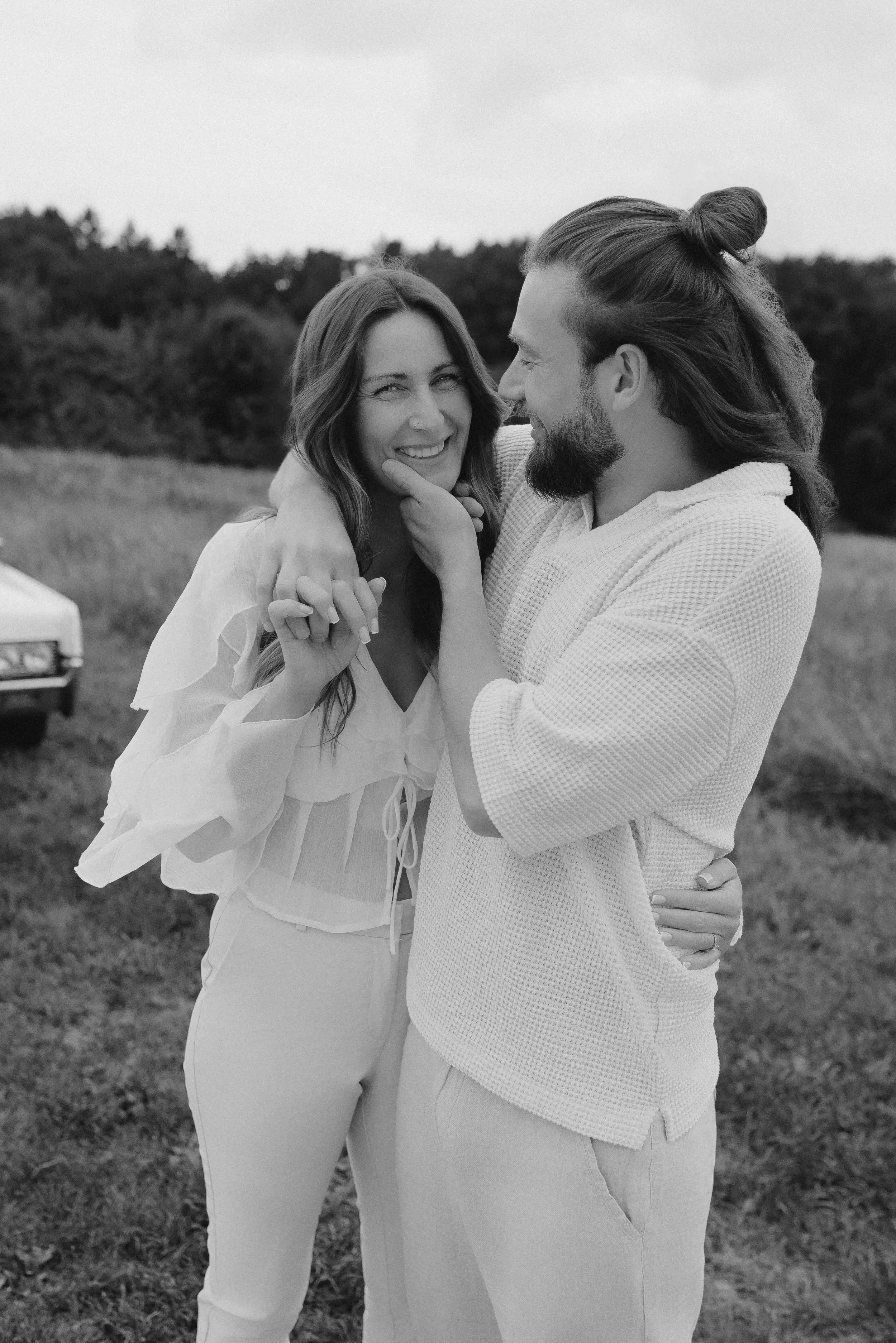 Ein glückliches Paar umarmt sich im Freien auf einer Wiese, lächelt und schaut sich liebevoll an, im Hintergrund Bäume und Wolken.
