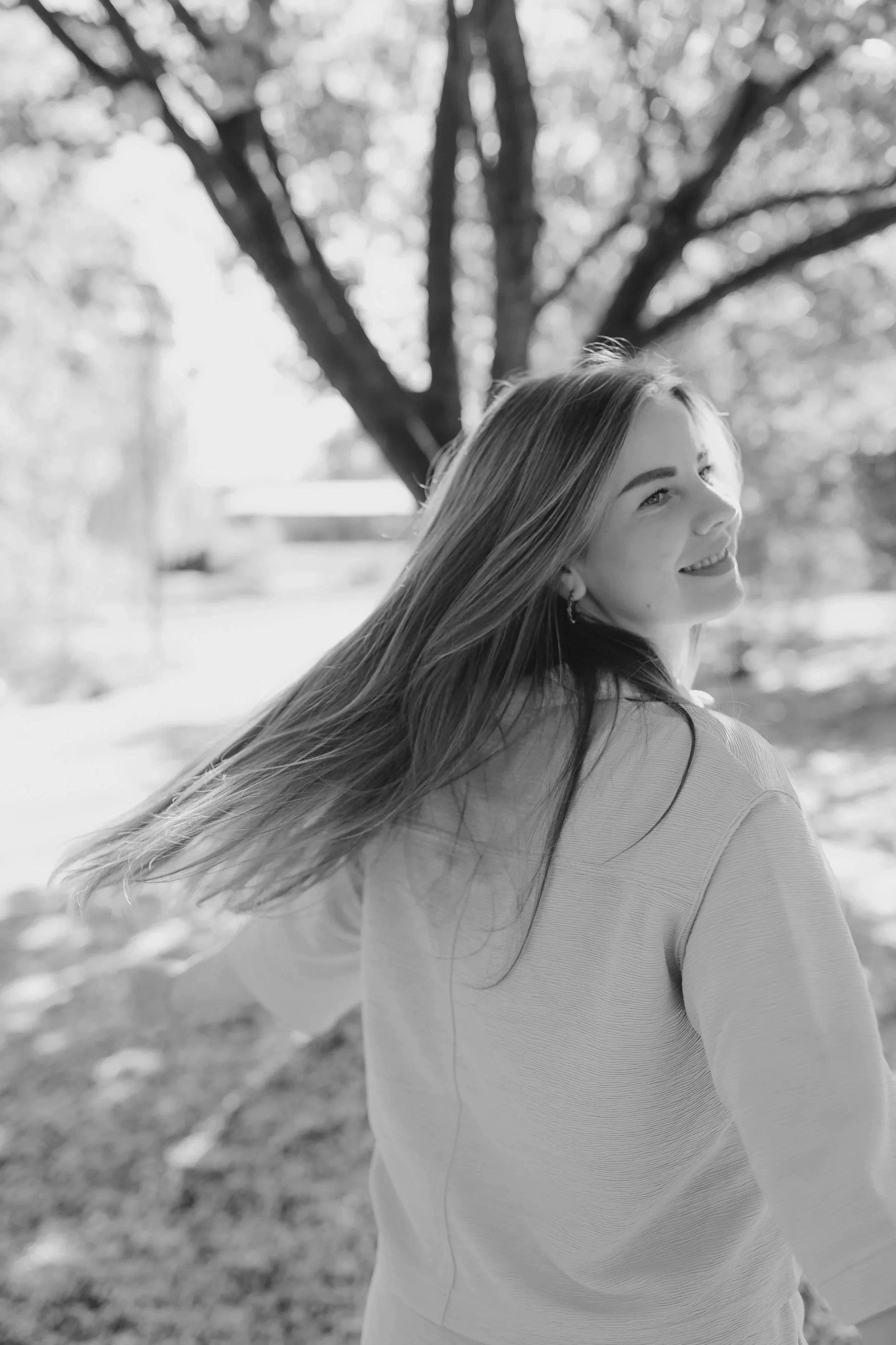 Schwarz-weiß-Fotografie eines lachenden Mädchens im Freien mit langer, wehender Haar, im Hintergrund Bäume.