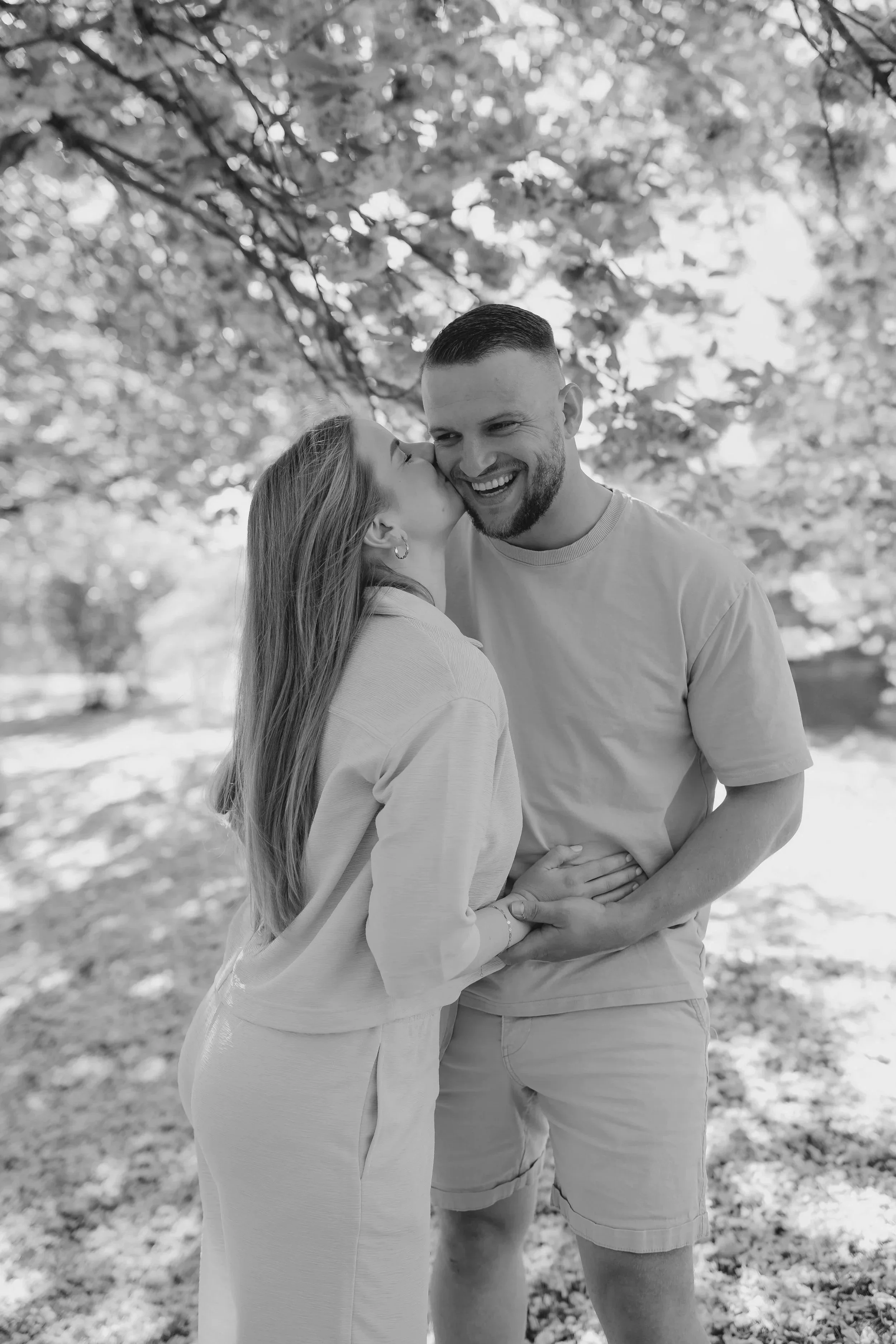 Ein Paar umarmt sich im Park, die Frau küsst den Mann auf die Wange, beide lachen, im Hintergrund Bäume, Schwarz-Weiß-Foto.