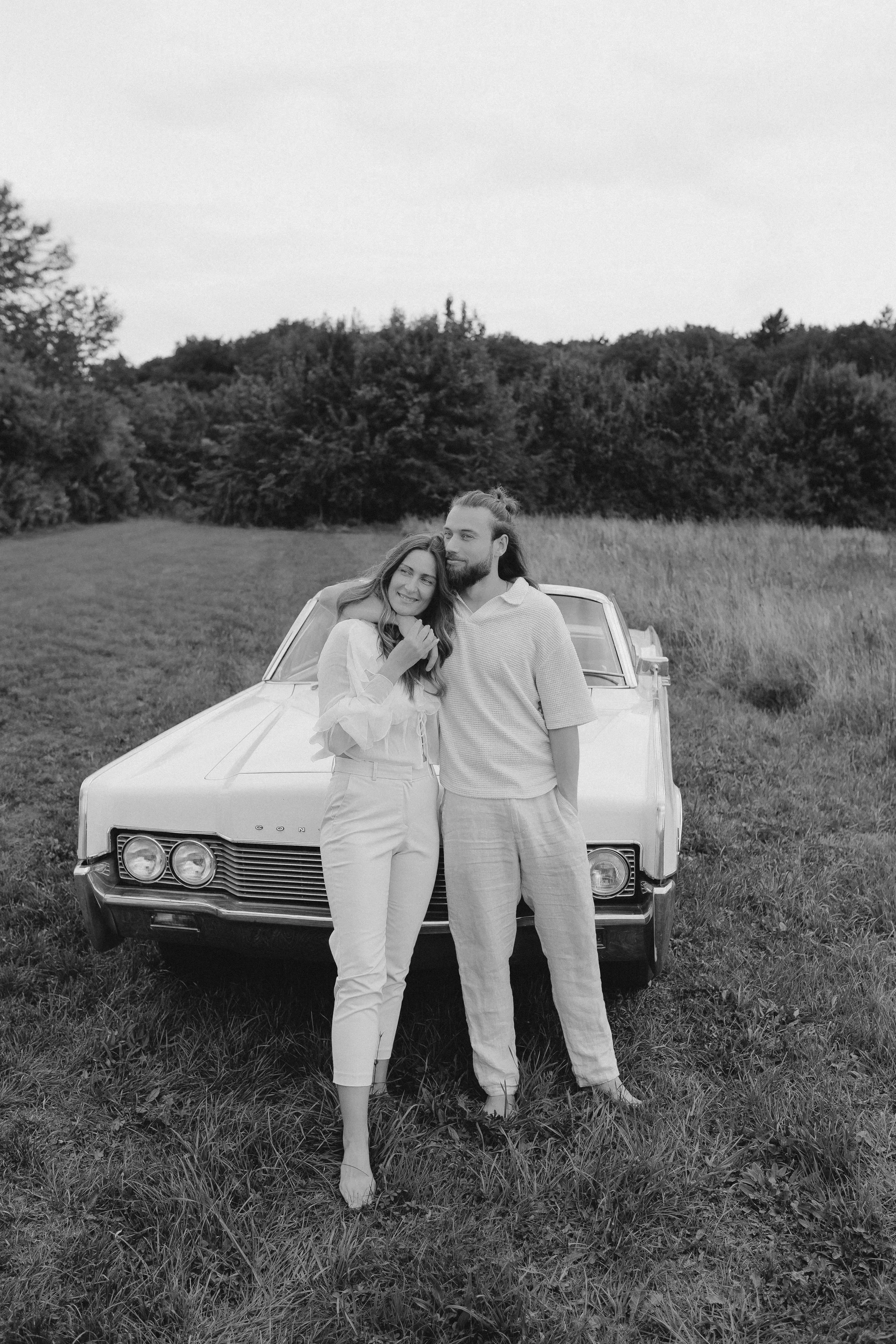 Ein Paar steht vor einem alten Auto auf einer Wiese, umgeben von Bäumen, fotografiert in Schwarz-Weiß.